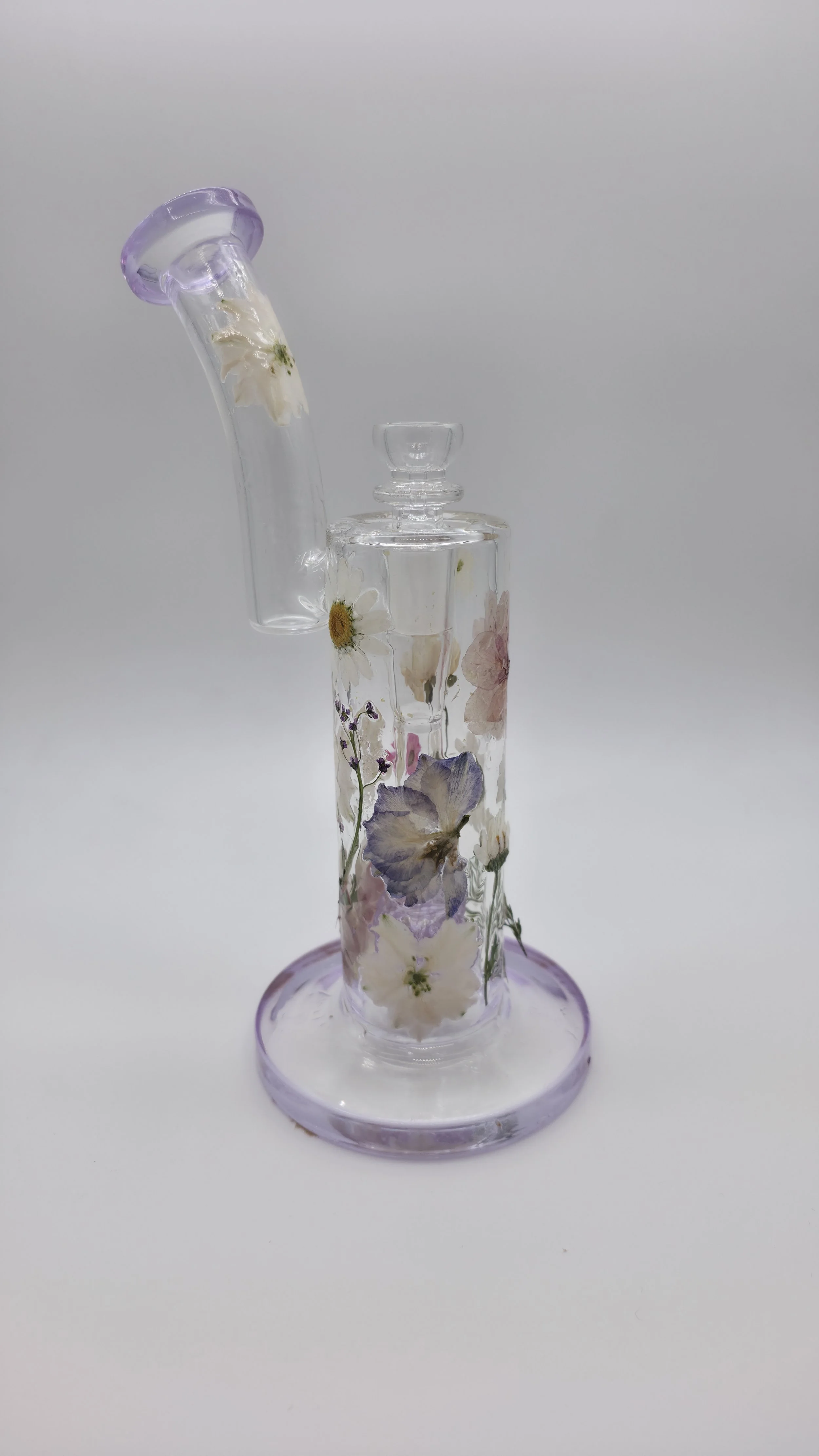 A glass bong decorated with pressed flower designs, with a daisy flower inside and dried flowers in the water chamber, set against a plain white background.