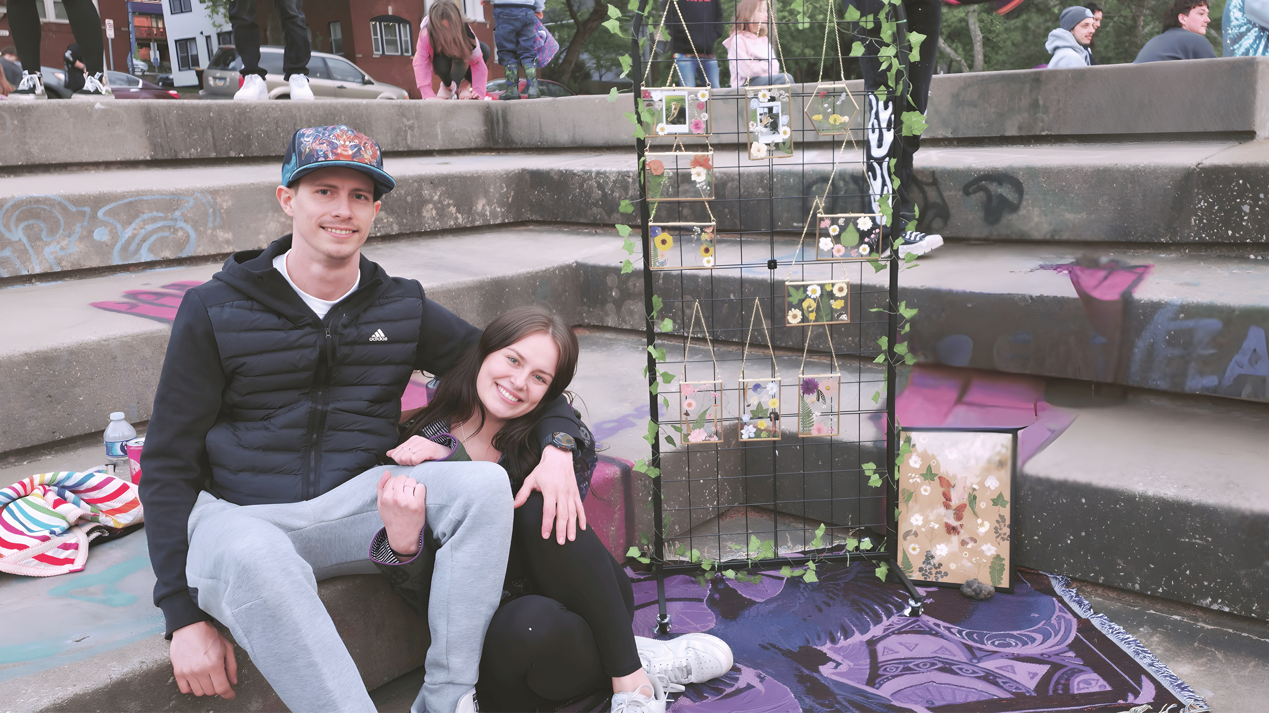 A young man and woman sitting together on steps at an outdoor event, smiling at the camera. There is a decorative display with picture frames and greenery behind them. Other people are visible in the background at the event.