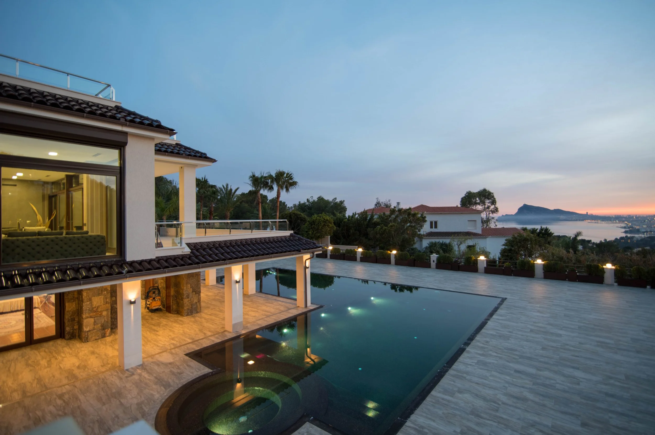 A modern house with large glass windows, a swimming pool, and a scenic view of trees, water, and mountains at sunset.