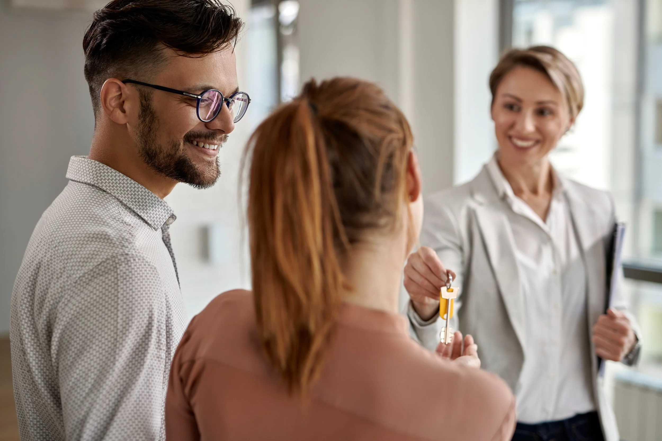 A man with glasses and a beard smiling and talking to women, one of whom is holding a key, in a bright modern office setting.