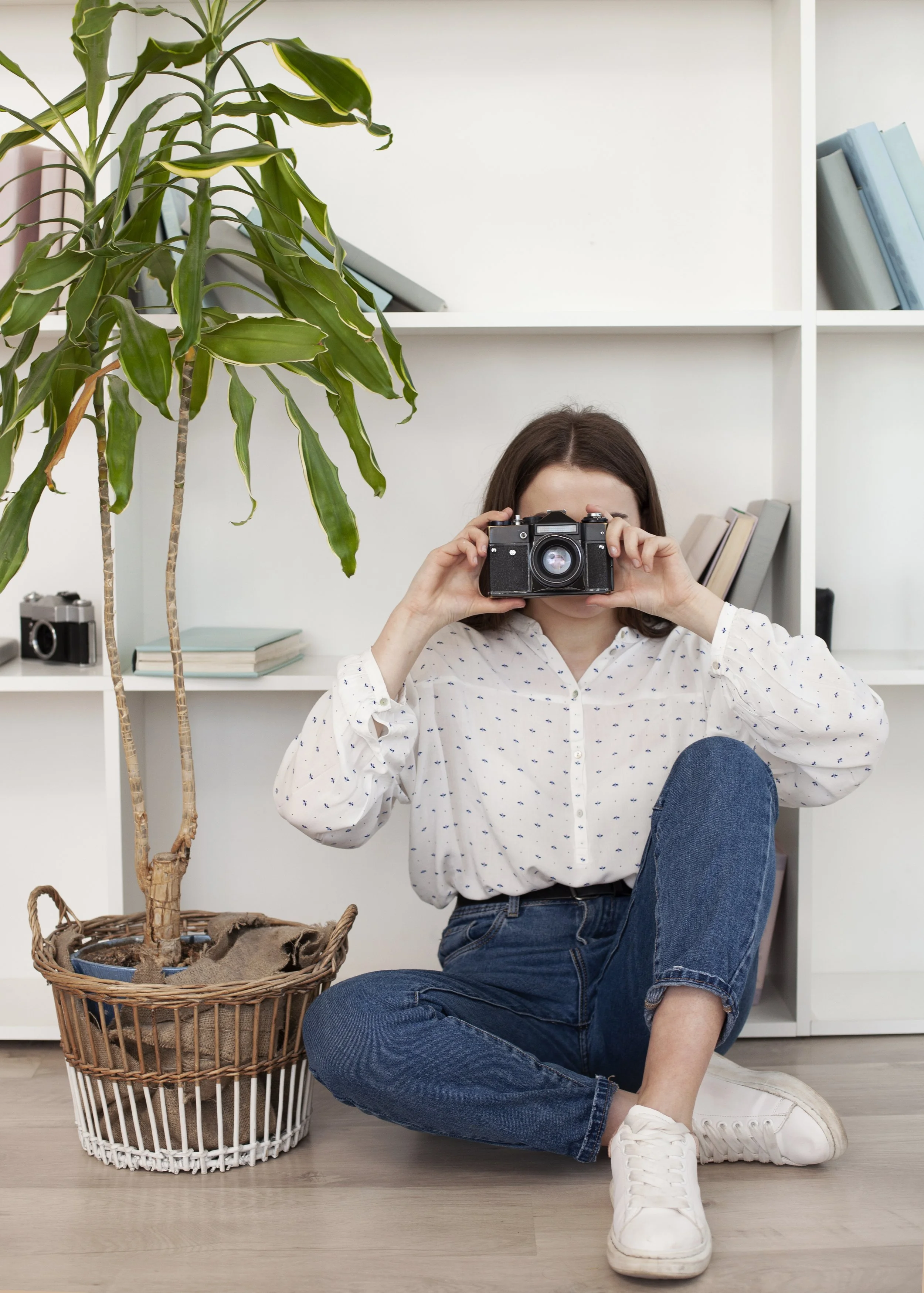 A woman sitting on the floor in front of a white bookshelf, holding a camera up to her face. There is a large potted plant in a wicker basket to her left, and books on the bookshelf behind her.