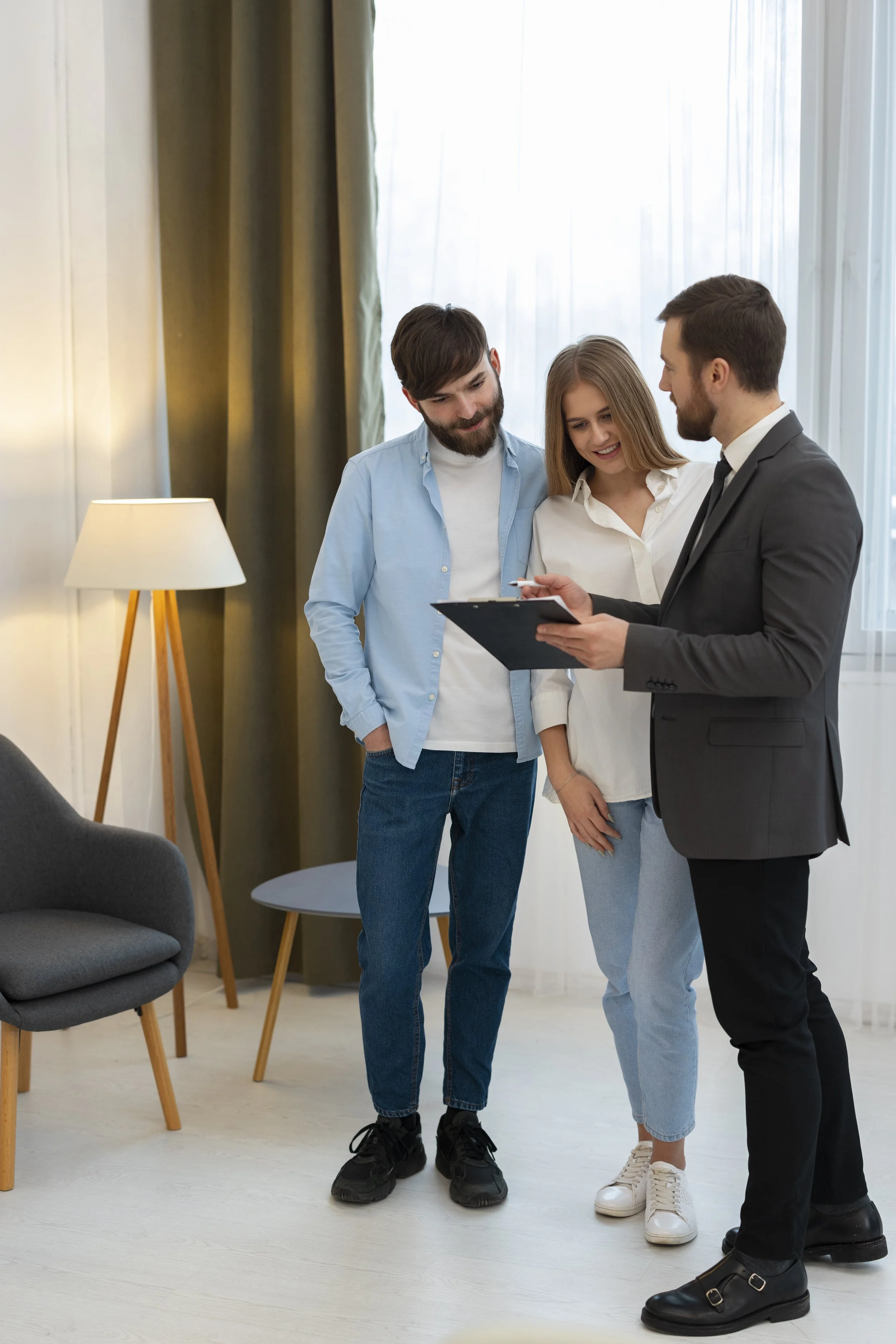 Three young adults, two men and one woman, are standing together in a well-lit room looking at a clipboard held by a man in a suit. They are smiling and appear engaged in a discussion.