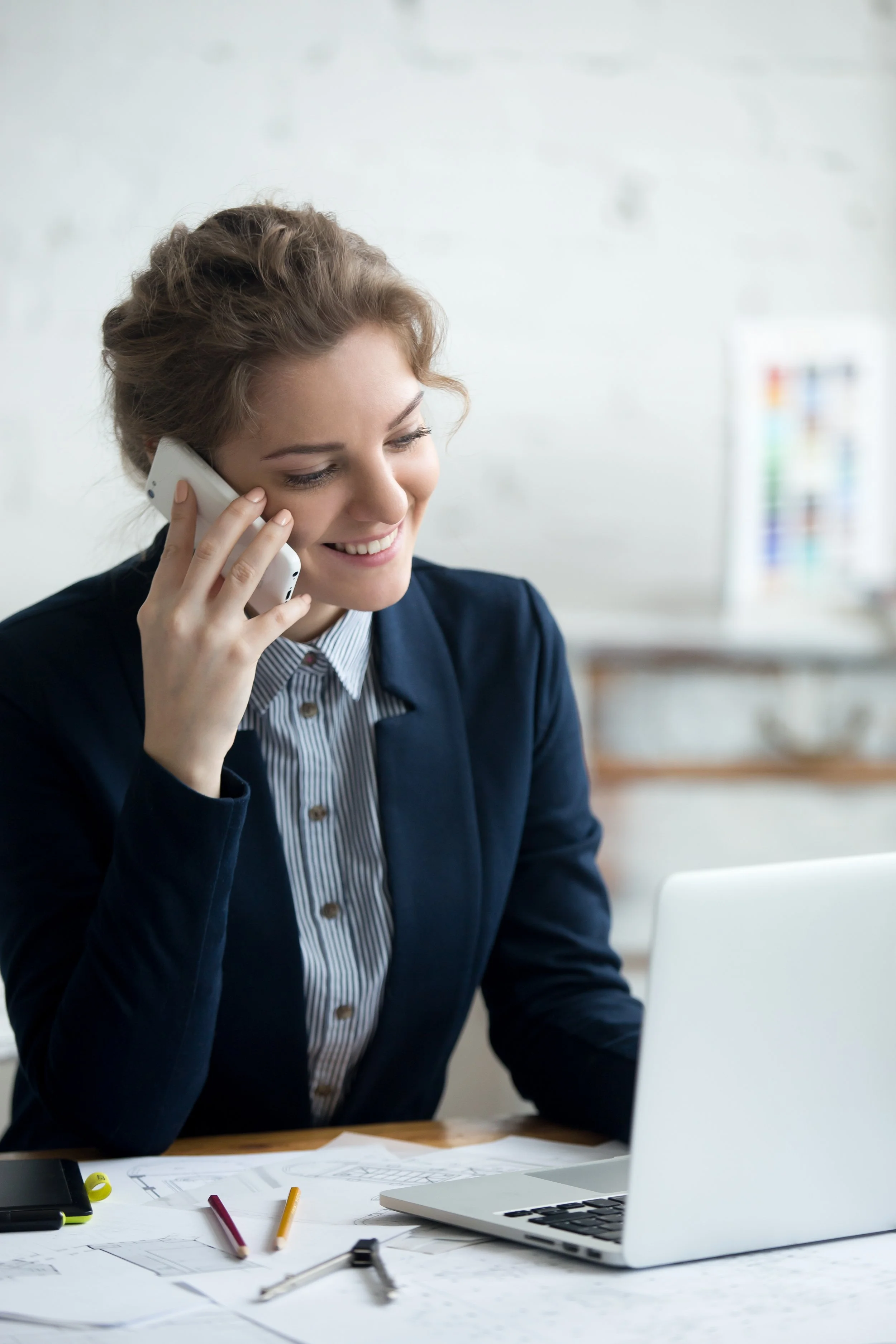 A young woman with brown hair smiling and talking on a mobile phone while working at a desk with various papers, colored pencils, and a laptop.