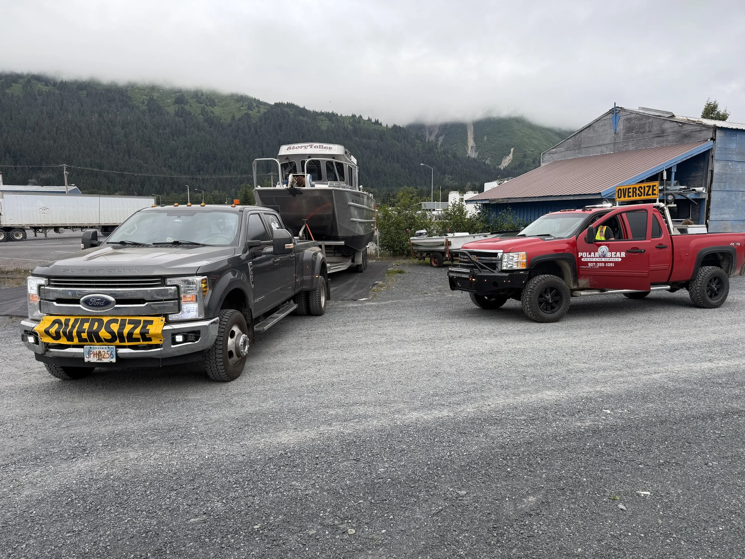 Two pickup trucks with oversized signs, one black and one red, parked on a gravel lot. A boat on a trailer is behind the black truck. Mountains and cloudy sky in the background.