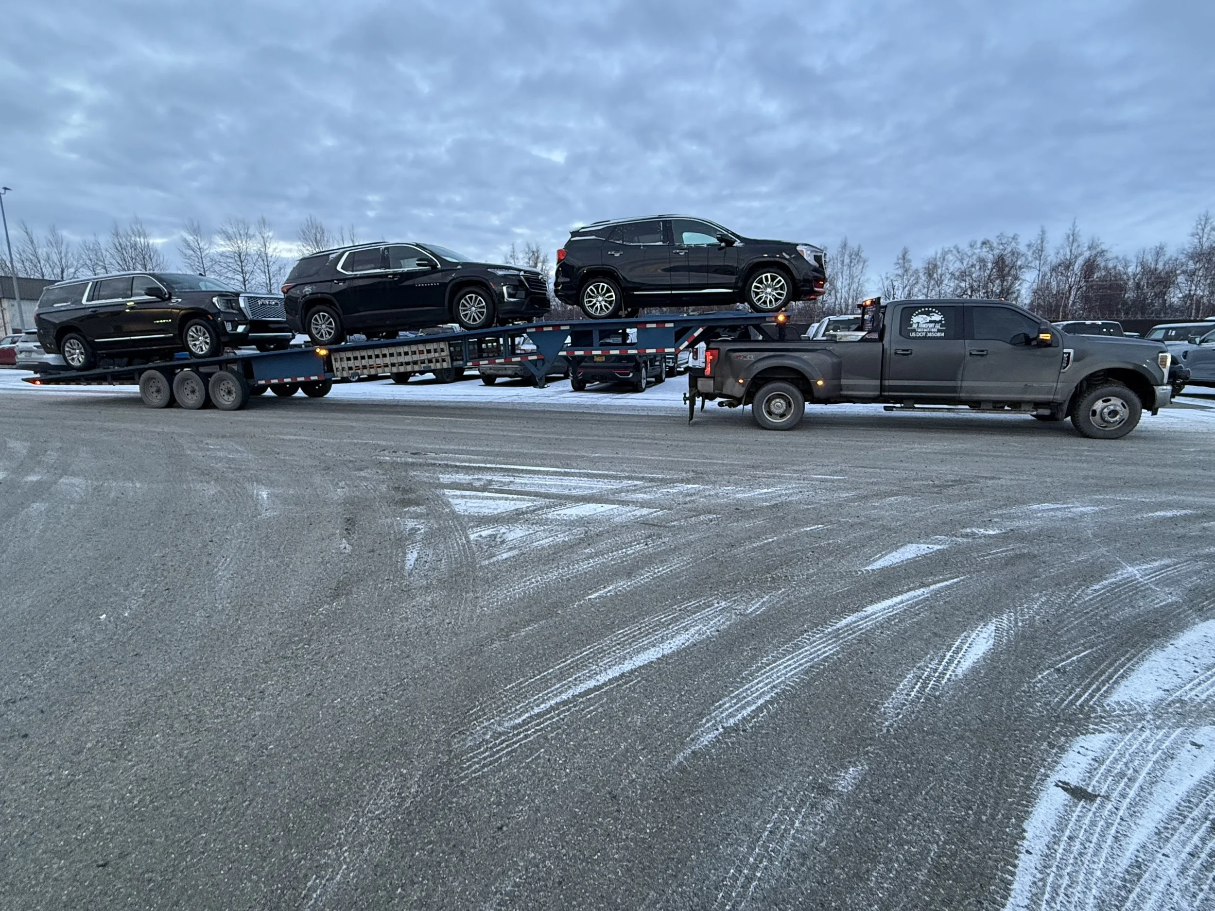 Black pickup truck transporting several black SUVs on a flatbed trailer in a snowy parking lot.