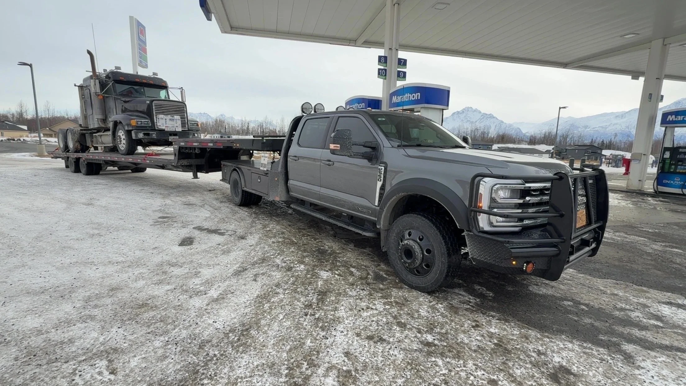 A tow truck at a gas station with snow on the ground, hauling a semi truck on its flatbed trailer, with mountains in the background.