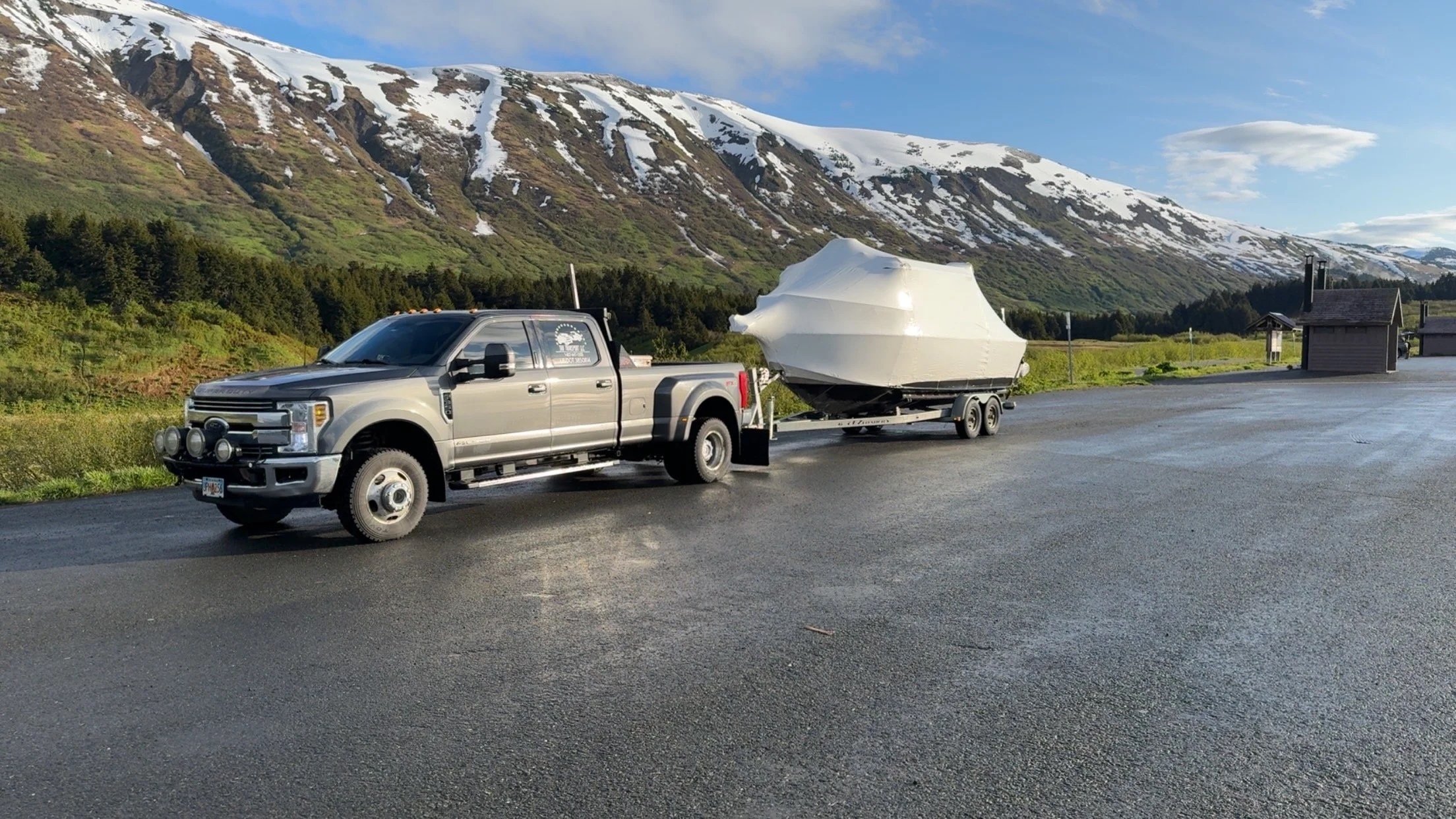 A large pickup truck pulling a boat on a trailer on a paved road with snow-capped mountains and a green landscape in the background.