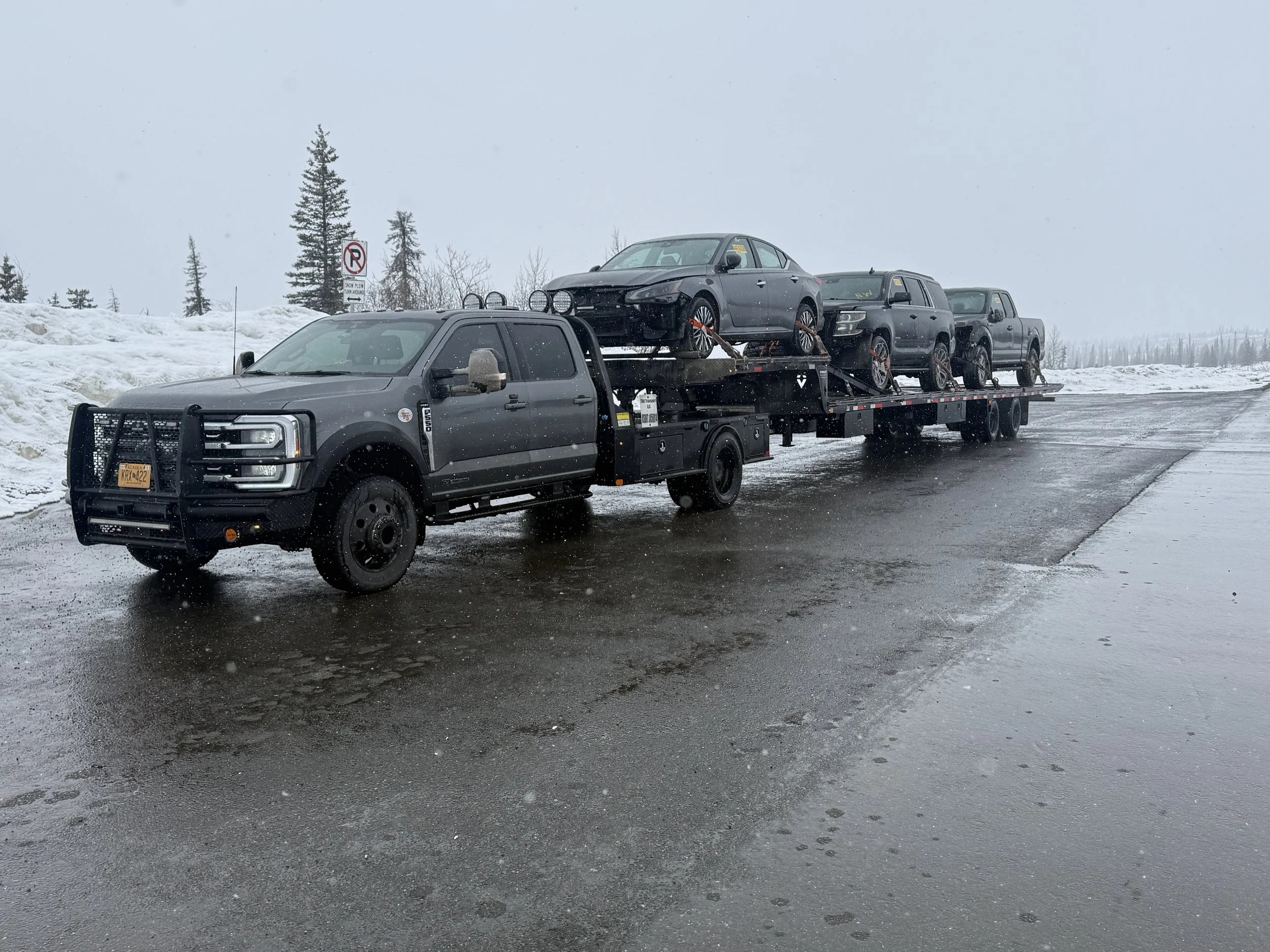 A black flatbed truck carrying four vehicles, including one with a front bumper and wheels removed, on a wet, snow-covered road during snowy weather.