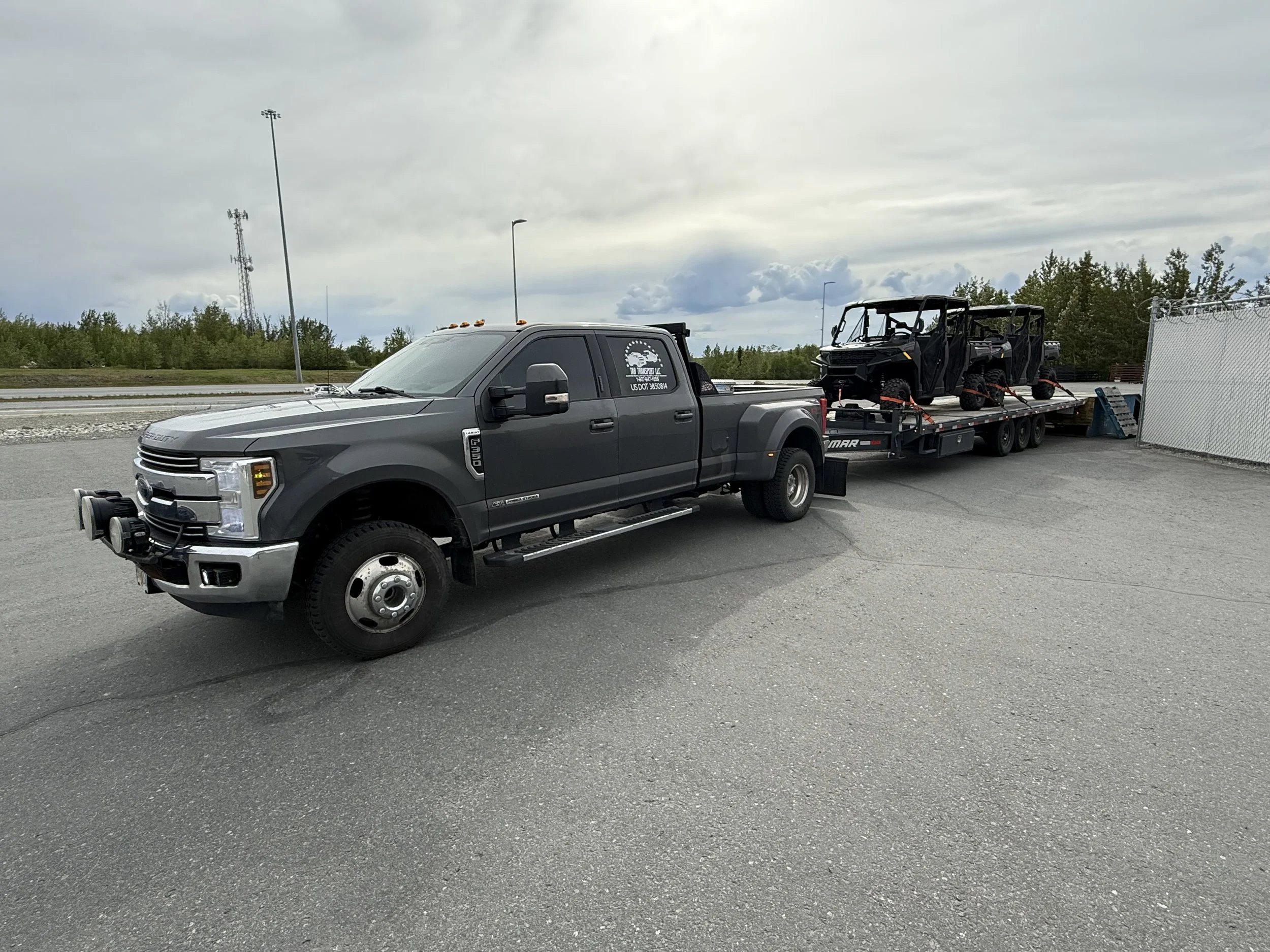 A black pickup truck hauling two vehicles on a flatbed trailer in a parking lot with cloudy skies and trees in the background.