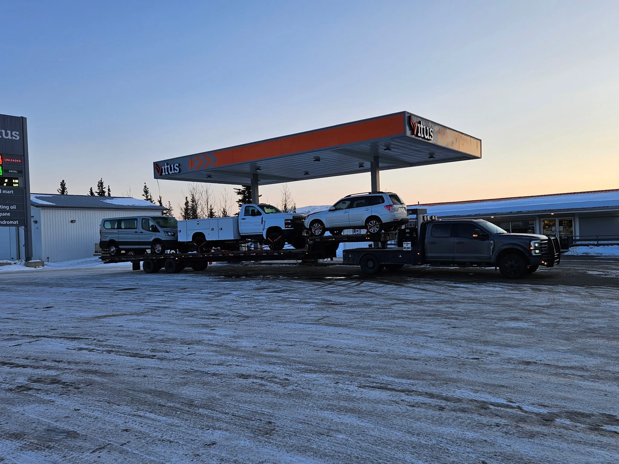 A transportation truck carrying multiple vehicles in a snow-covered parking lot at a vitus station during dusk.