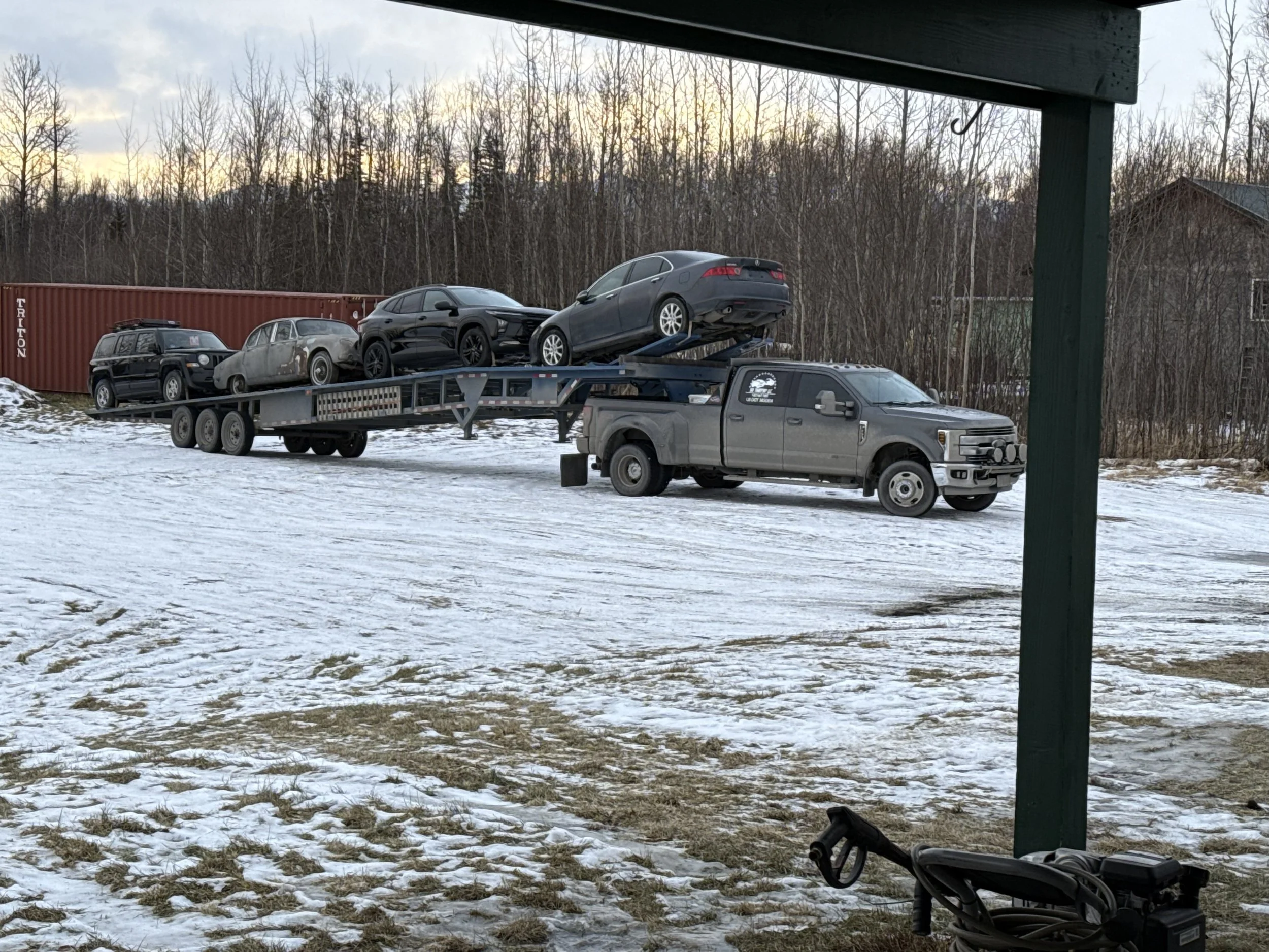 A flatbed truck hauling several cars on a snowy landscape with trees in the background, partly icy and snowy ground, and a wooden structure in the foreground.