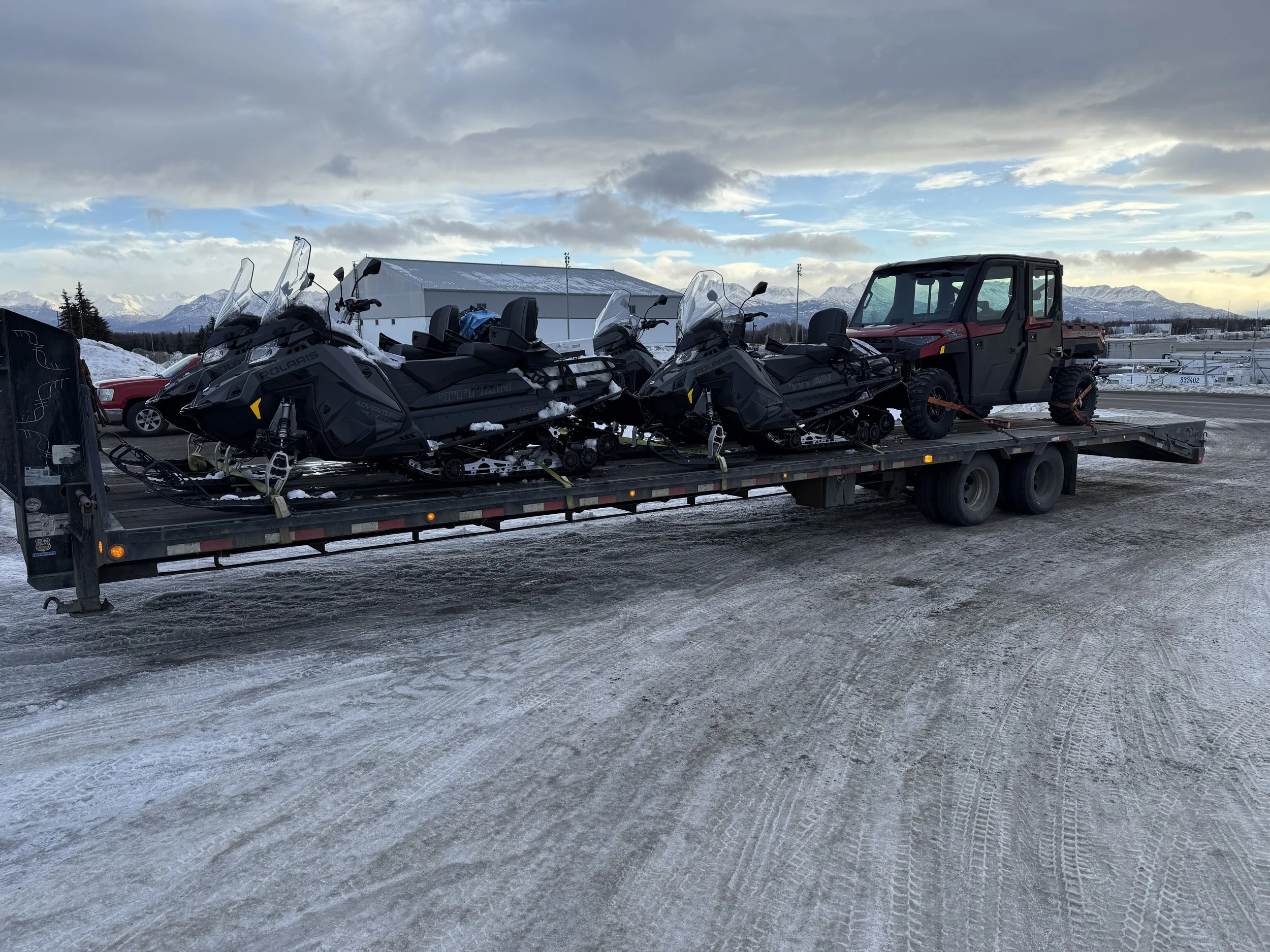 A flatbed trailer carrying two snowmobiles and a utility ATV in a snowy parking lot with mountains in the background.