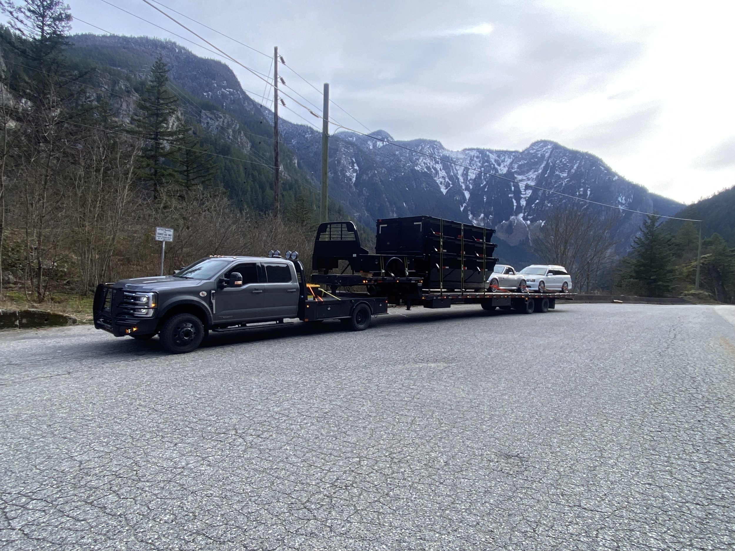 A flatbed truck carrying two white vehicles and a black cargo container, parked on a mountain road with snow-capped mountains and trees in the background.