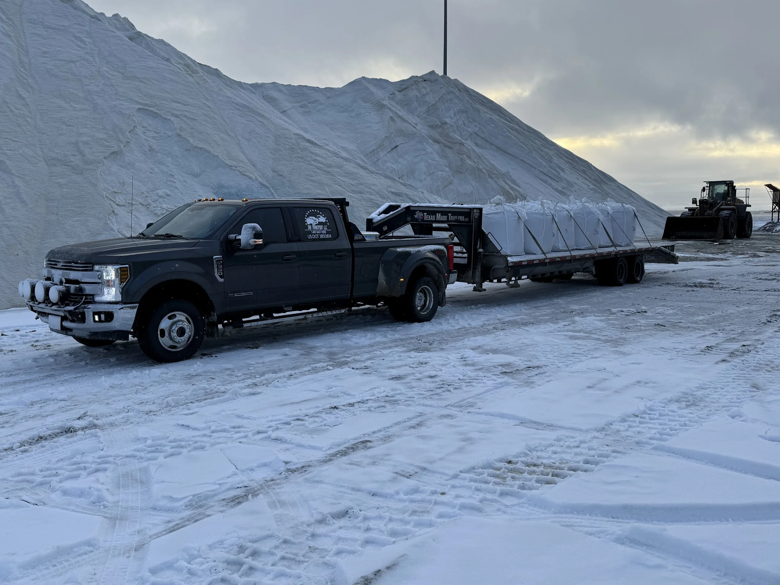 A black pickup truck with a trailer is parked on snow-covered ground near large white salt piles, with a bulldozer in the background on a cloudy day.