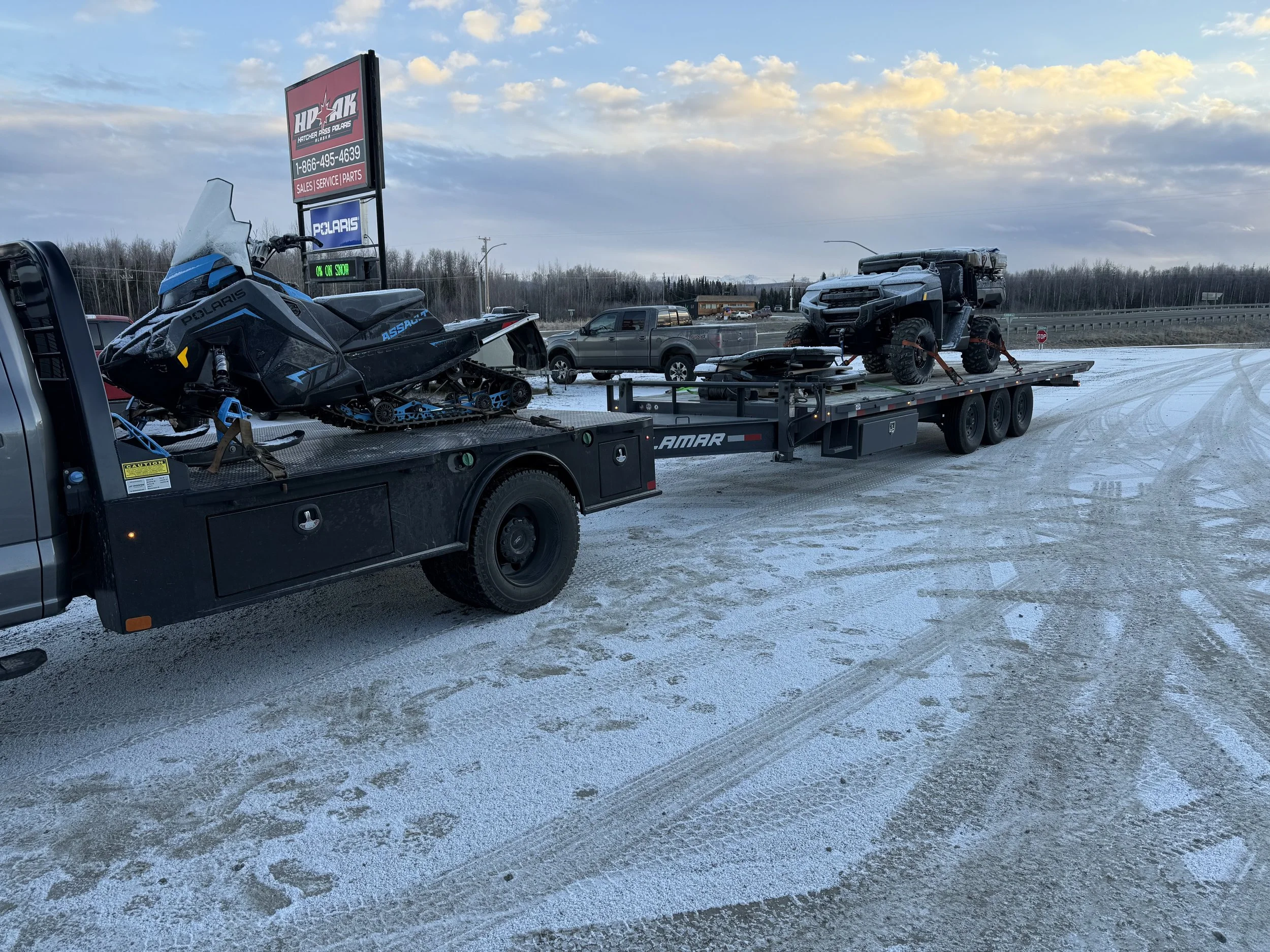 A flatbed truck transporting a snowmobile and an off-road vehicle on a snowy, icy roadside with a dealership sign in the background.