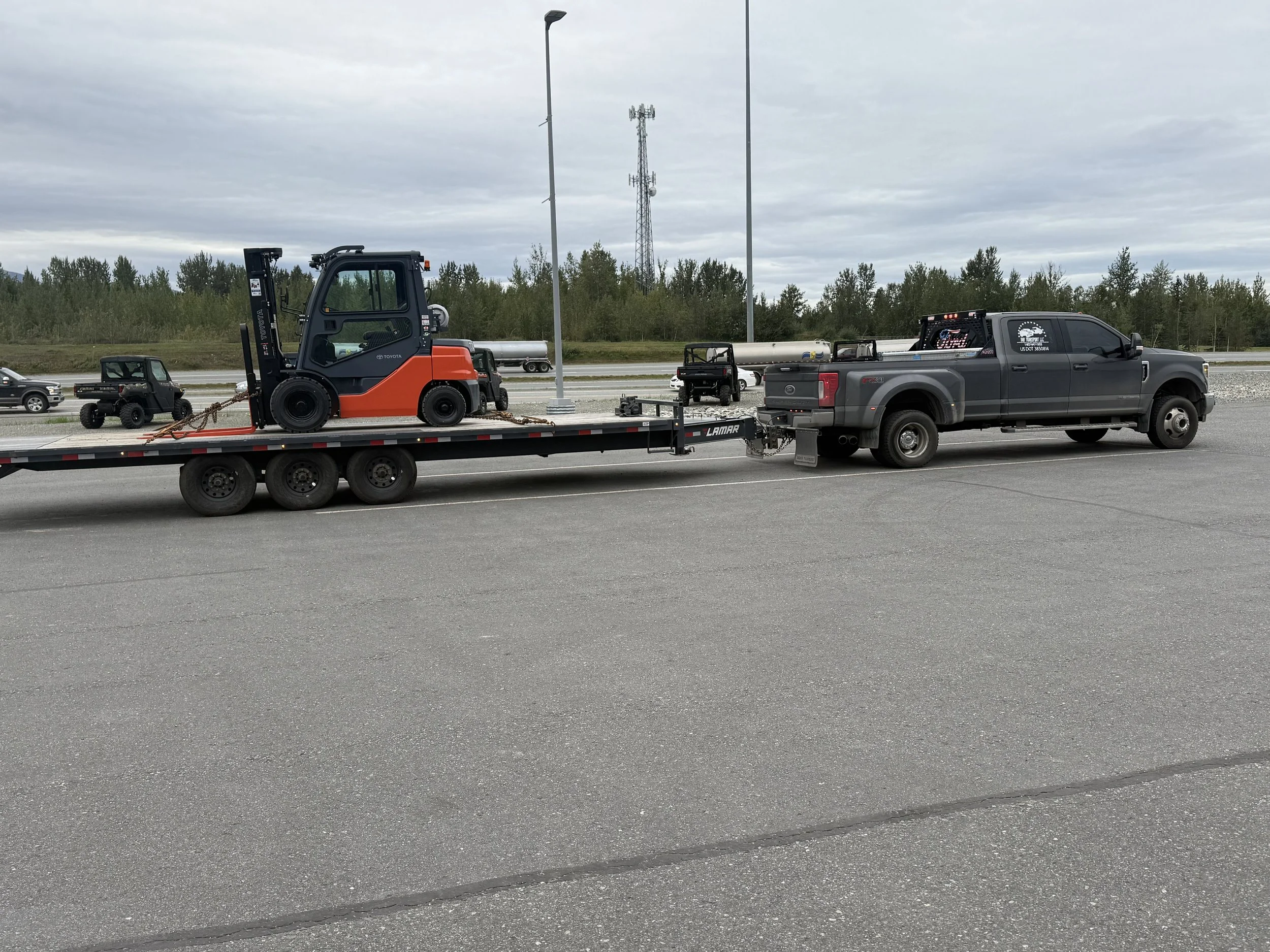 A black pickup truck towing a flatbed trailer with a small orange and black forklift on it.