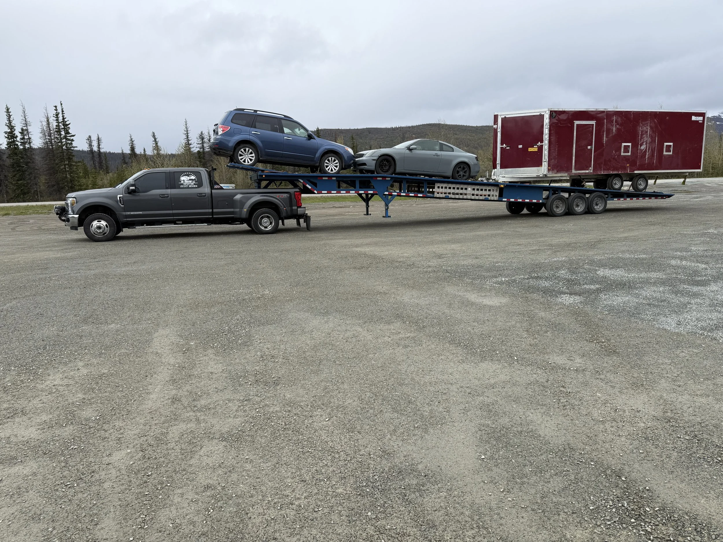 A black flatbed tow truck transporting two cars and a large red trailer in an open area with a cloudy sky and distant trees.
