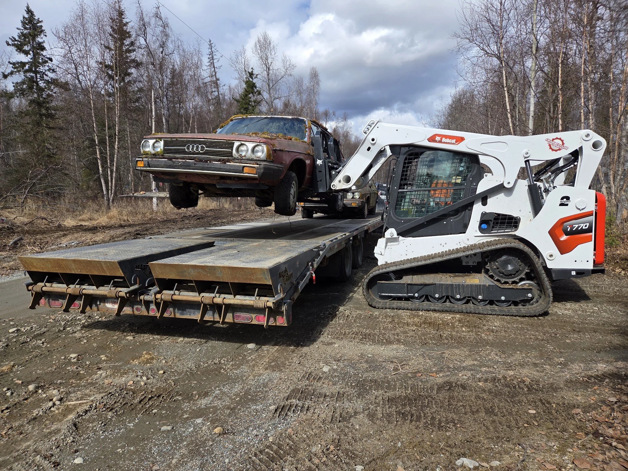 A compact loader lifting a rusted vintage Audi car onto a flatbed truck in a forested area with leafless trees and a cloudy sky.