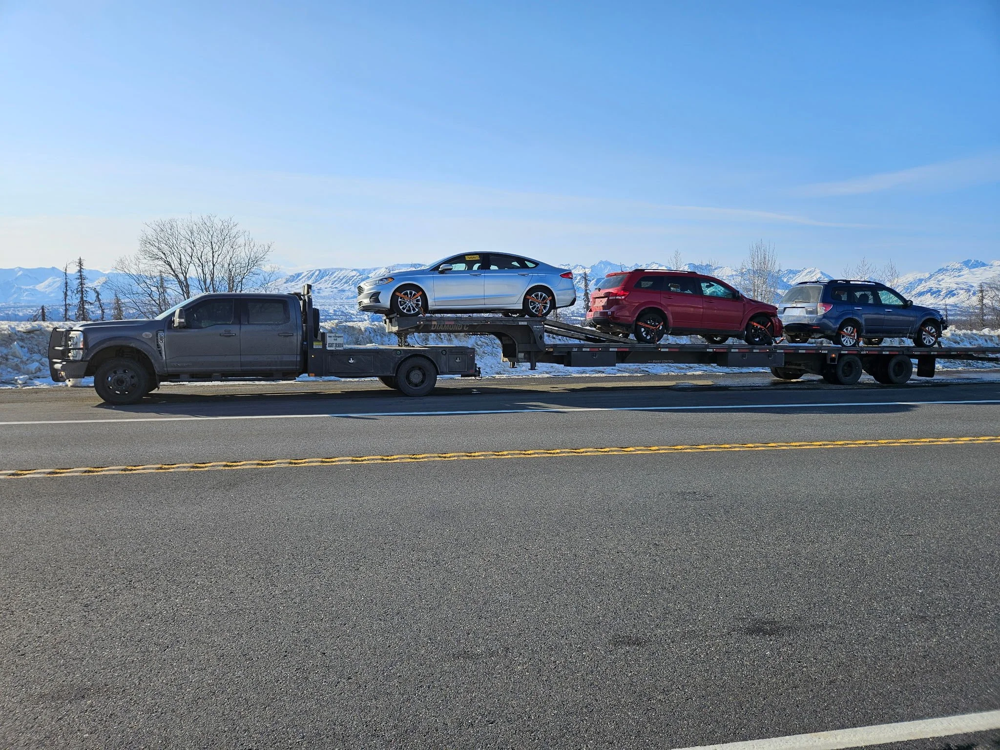 Black flatbed tow truck transporting three passenger vehicles on a highway with snow-covered mountains in the background.