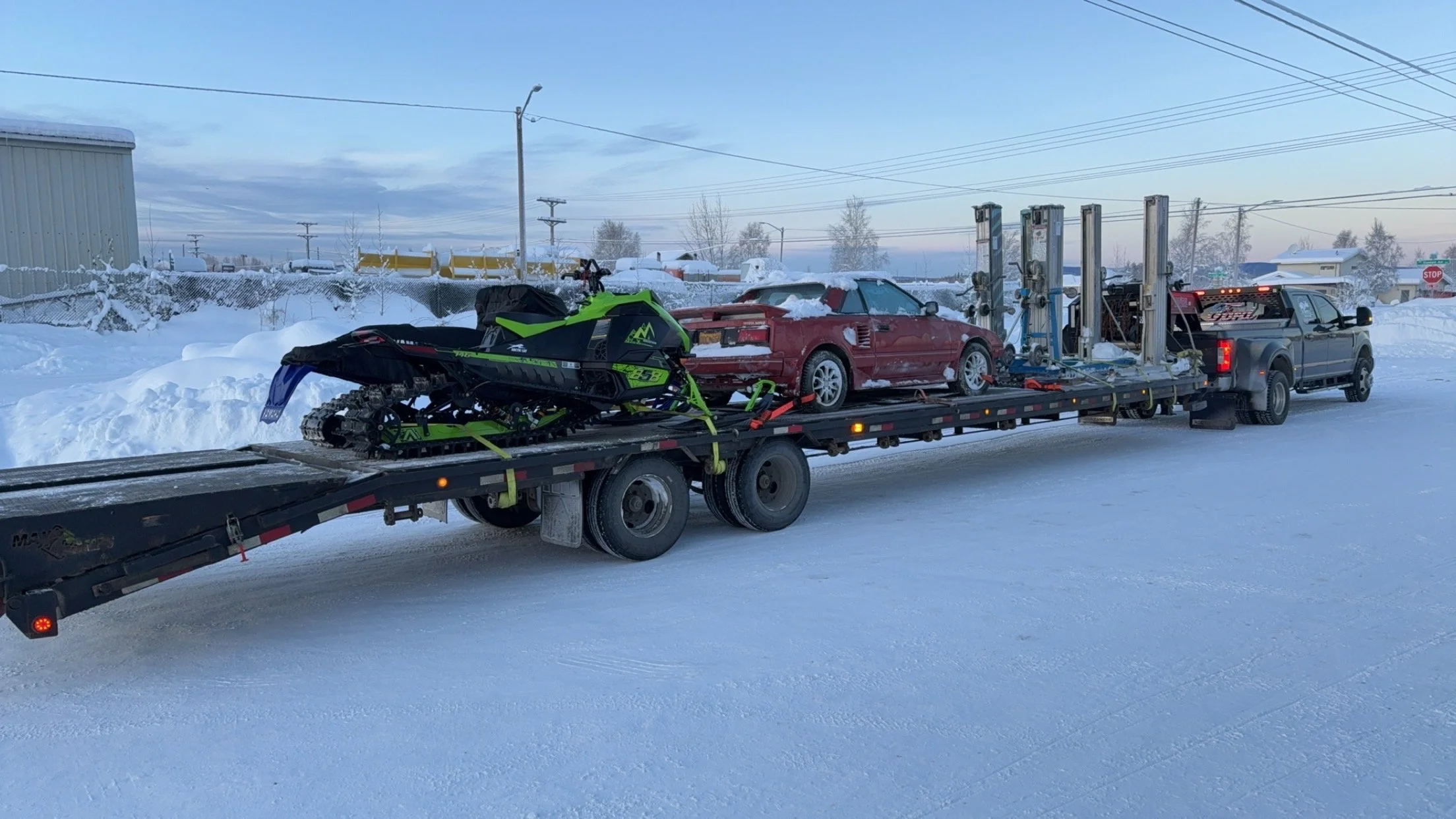 A flatbed tow truck carrying a red sports car covered in snow and a snowmobile on a snowy road in a winter landscape.