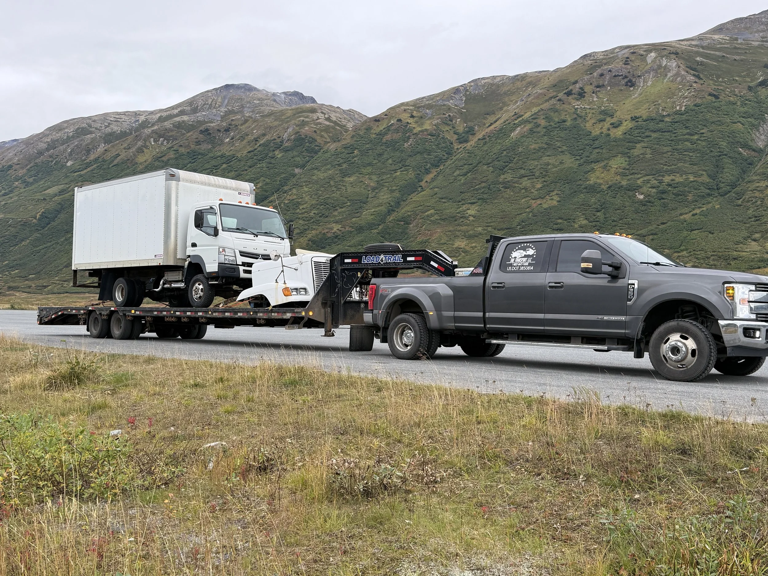 A black pickup truck towing a flatbed trailer with a white utility truck and a semi-truck cab on it, on a rural road with green mountains in the background.