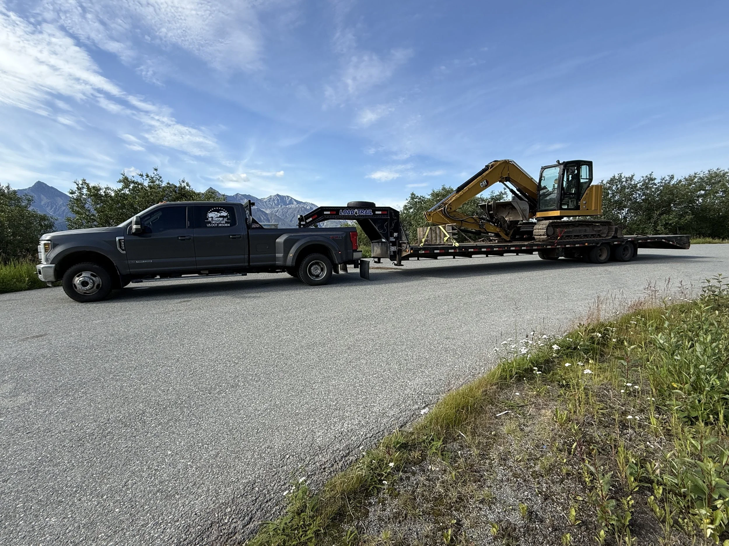 A black flatbed truck carrying a small yellow excavator with mountains in the background and a partly cloudy sky.