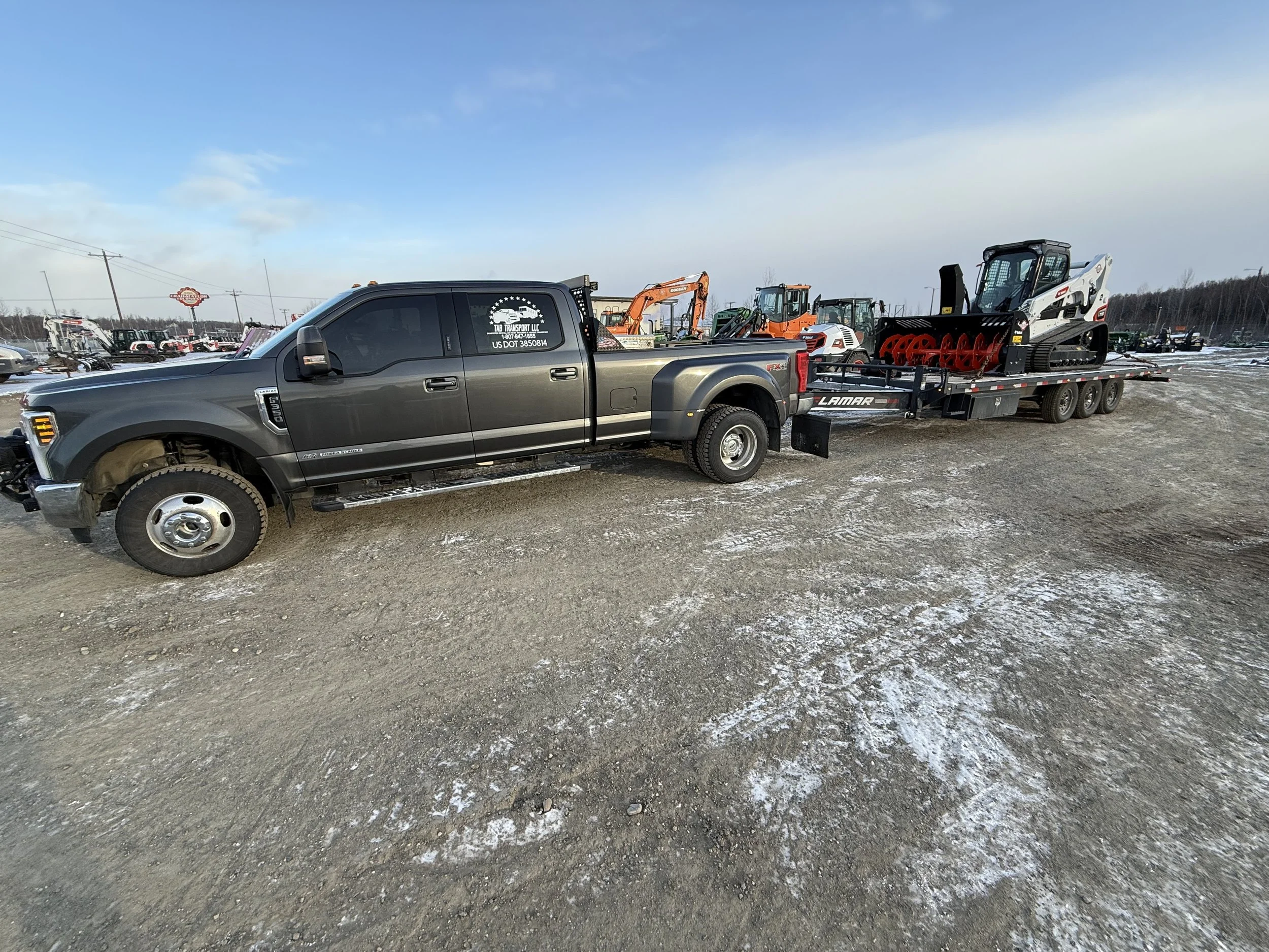 A black pickup truck with a flatbed trailer attached, carrying construction equipment including a skid steer loader and a compact excavator, parked on a gravel lot with a partly cloudy sky.