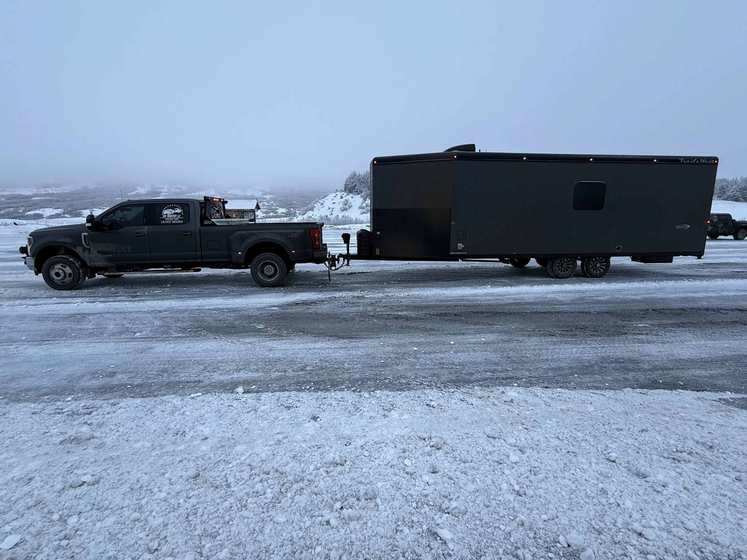 Black pickup truck pulling a large black trailer on a snow-covered road in a cold, foggy landscape with hills and trees in the background.