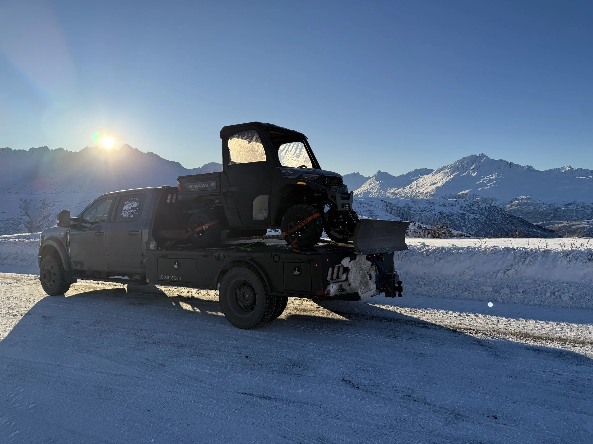 A black truck carrying a snow groomer on a snowy road with snow-covered mountains in the background during sunset.