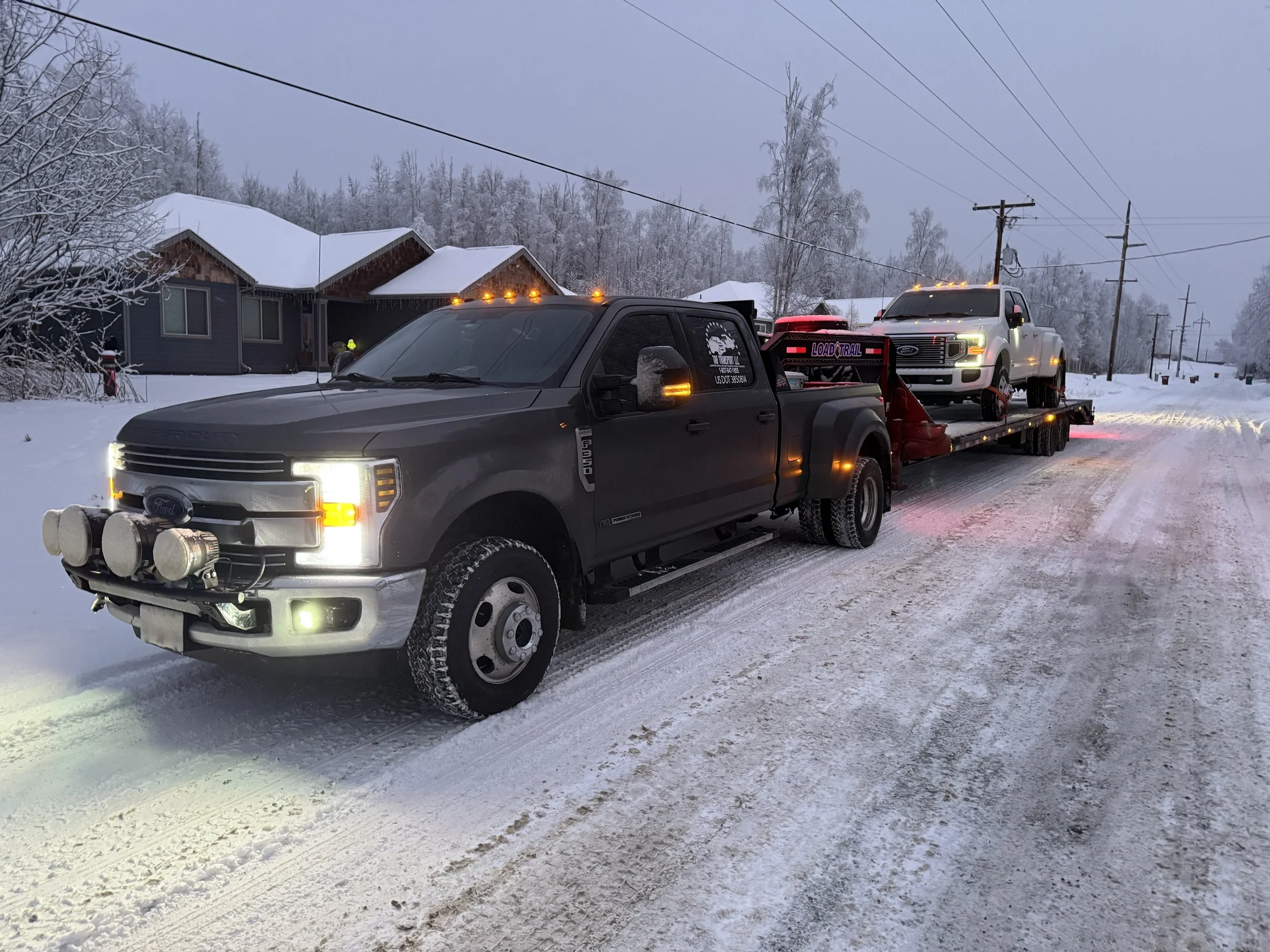 A dark gray Ford truck towing a flatbed trailer carrying a white pickup truck on a snowy road in a residential area with snow-covered houses and trees in the background.