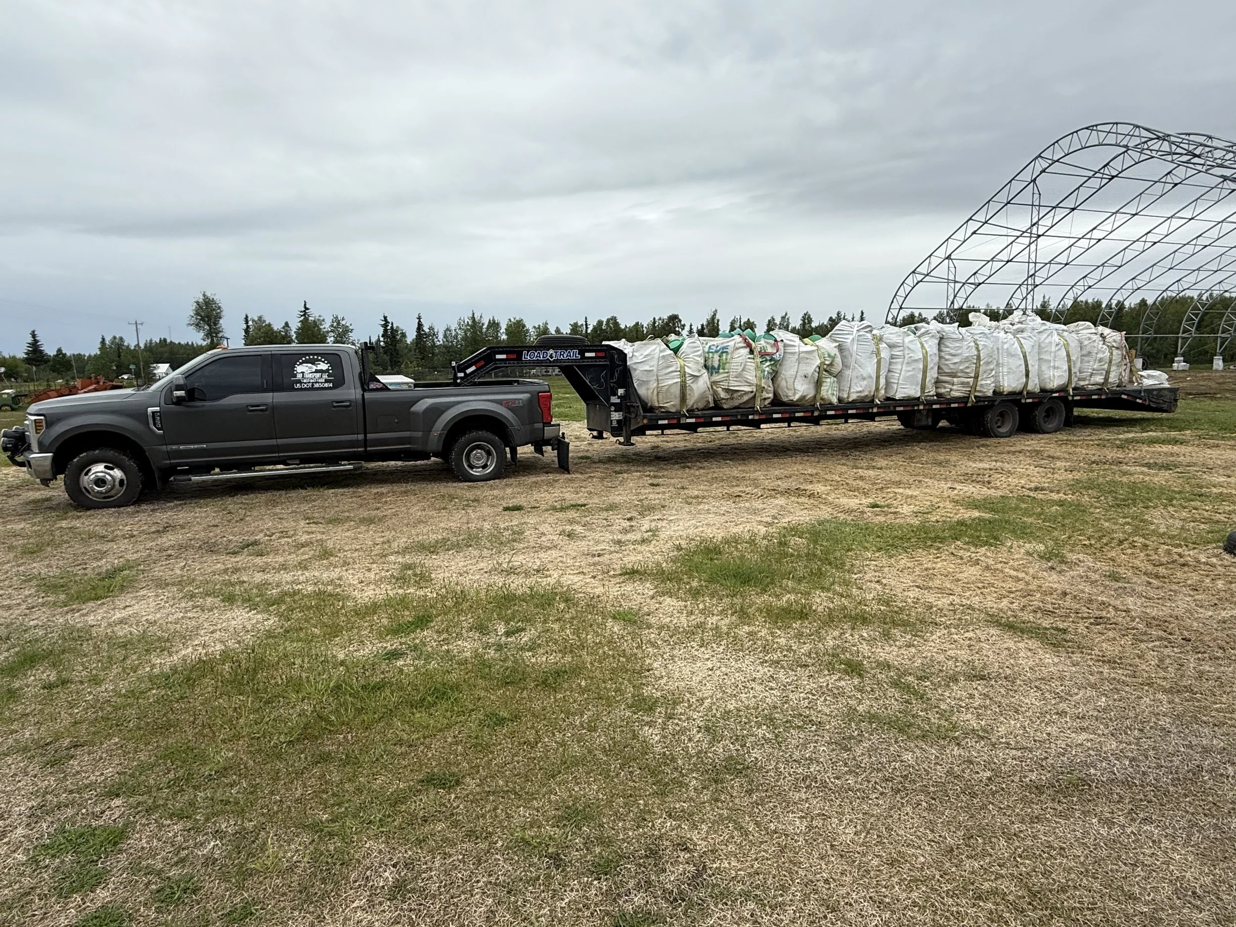 A large black pickup truck pulling a flatbed trailer loaded with white bags, parked on a grassy field under a cloudy sky.