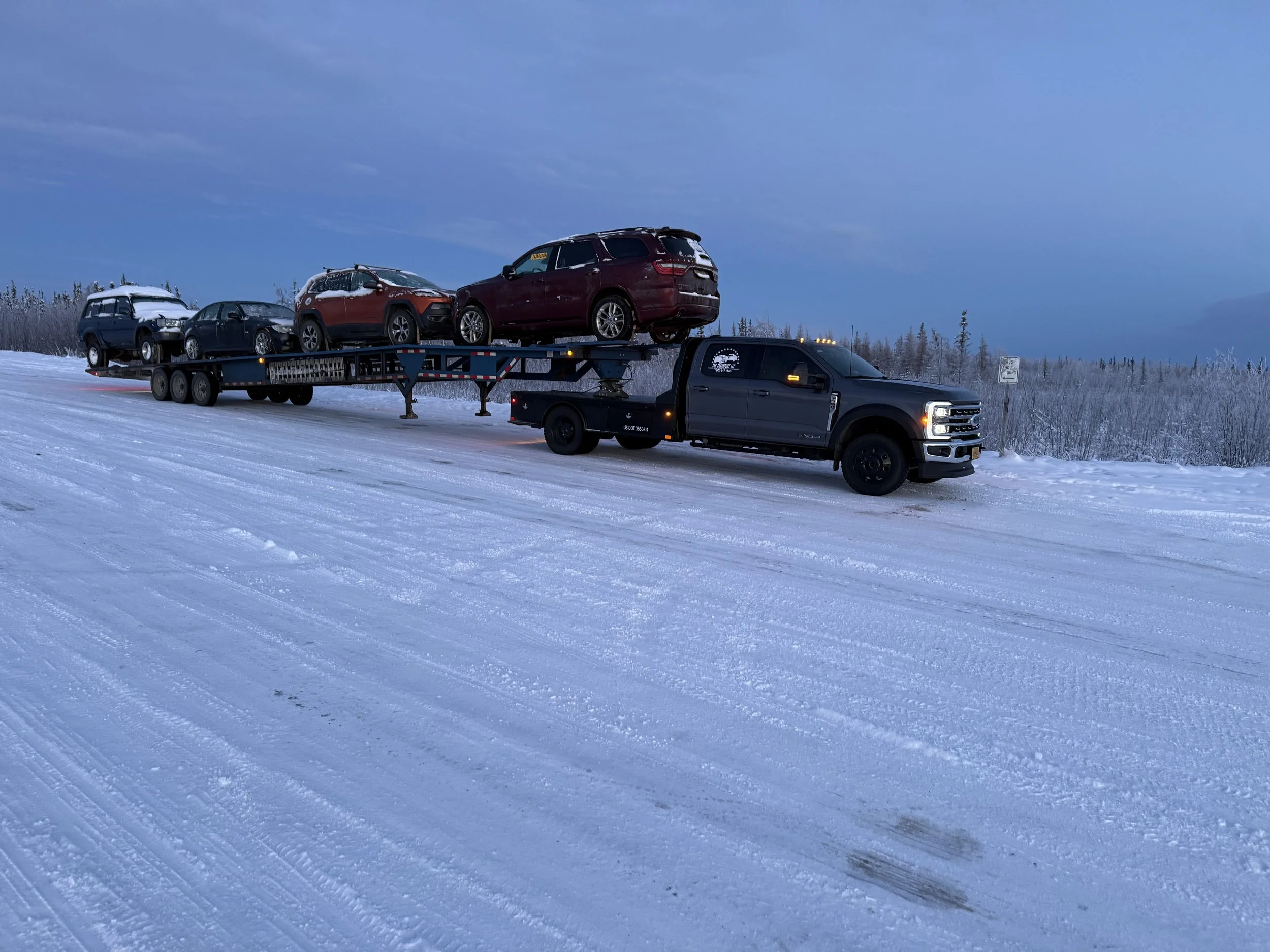 Black pickup truck towing a car carrier with four cars on a snowy road.