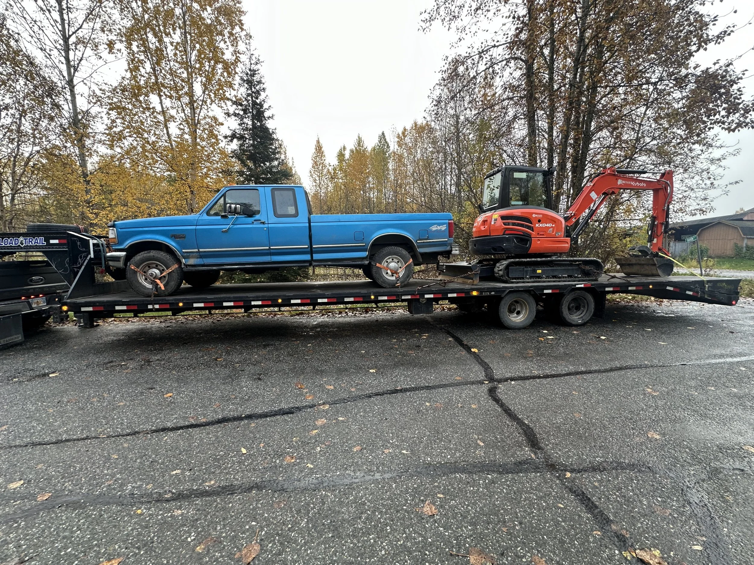 A blue pickup truck and a small red excavator loaded on a flatbed trailer parked on a wet paved surface, with trees and a house in the background.