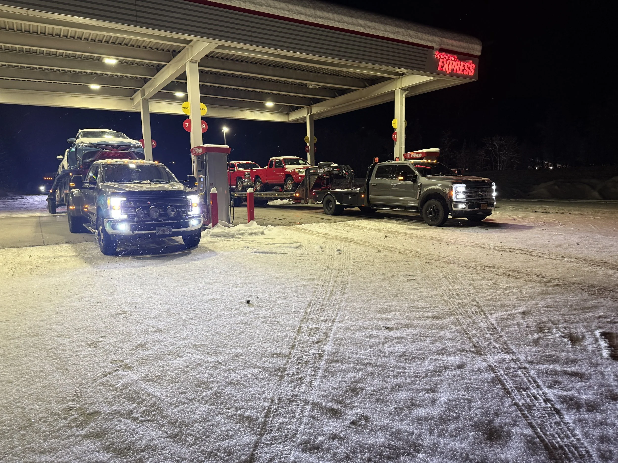 Snow-covered fuel station at night with multiple trucks parked, including a black truck carrying a car, and a red and black truck with additional vehicles on the servicing lot. Neon red 'Speedway Express' sign above.