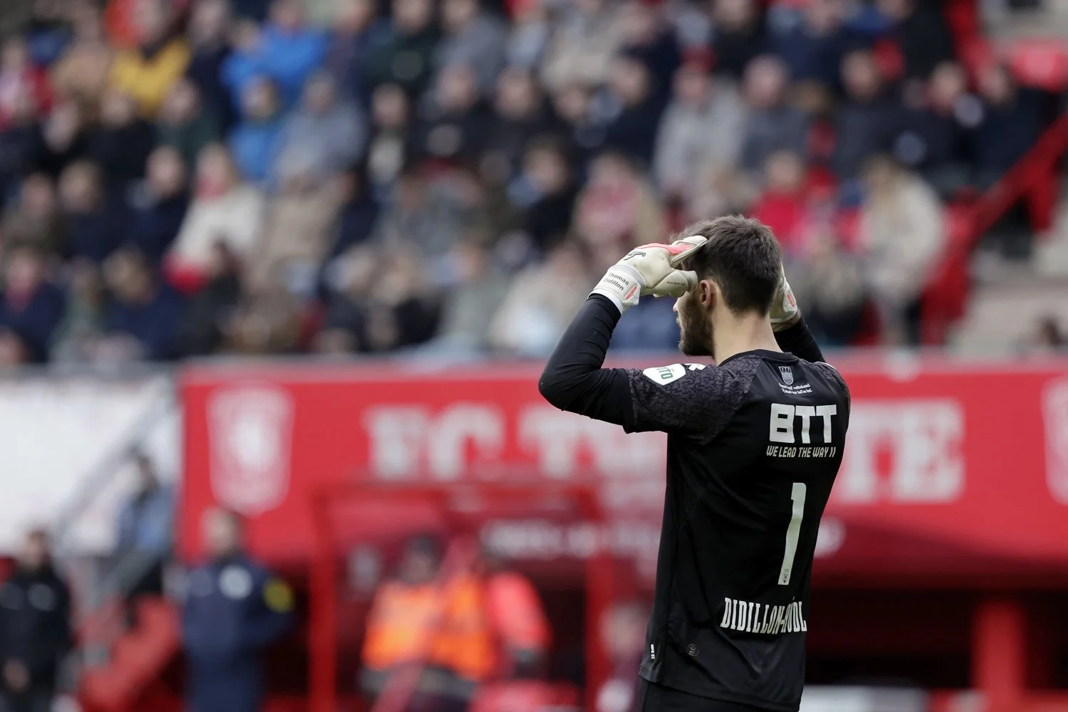 Un gardien de but de football portant un maillot noir avec le nom DIDILLON-HÖDL au dos, tenant ses mains sur sa tête lors d'un match, avec un public en arrière-plan.