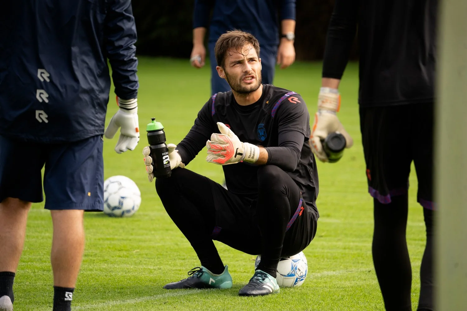 Thomas Didillon-Hödl assis sur le gazon lors d'un entraînement, entouré d'autres joueurs, tenant une bouteille d'eau, devant un ballon de football. Il semble expliquer quelque chose.