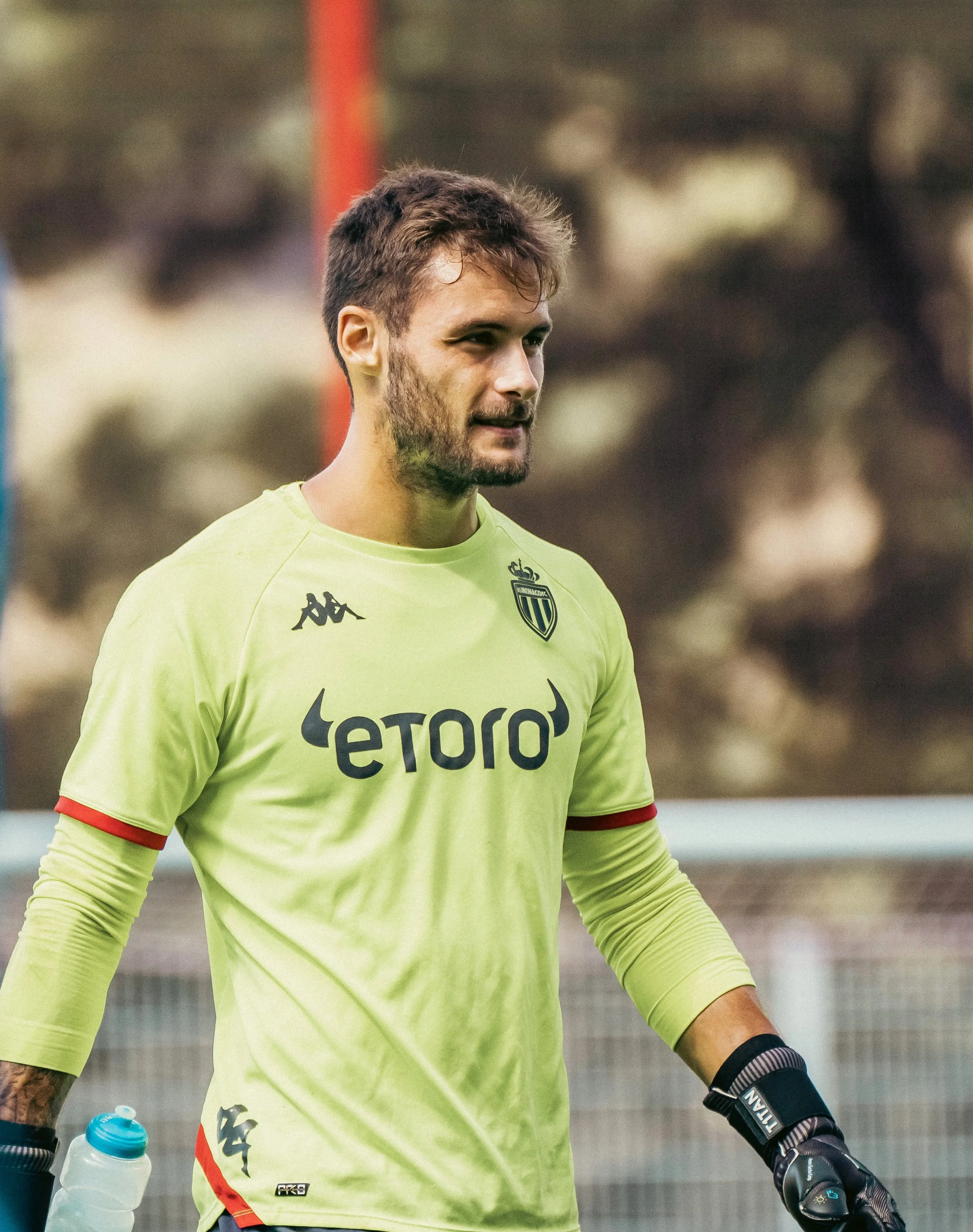 Un joueur de football en train de jouer sur le terrain. Il porte un maillot jaune avec le logo de la team et il tient une bouteille d'eau dans une main.