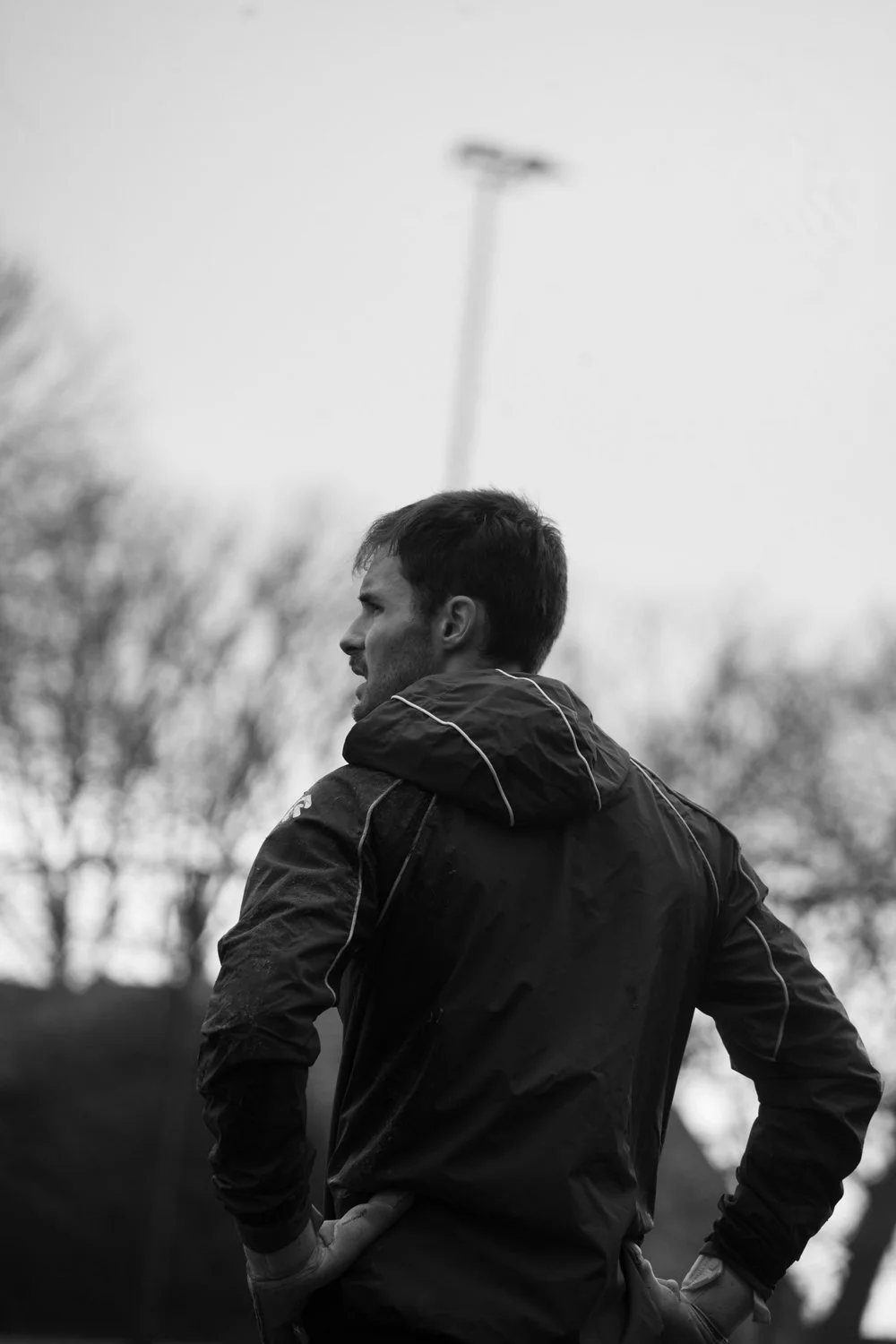 Homme en veste foncée avec capuche, en regardant vers la gauche, debout à l'extérieur avec des arbres et un lampadaire flous en arrière-plan, en noir et blanc.