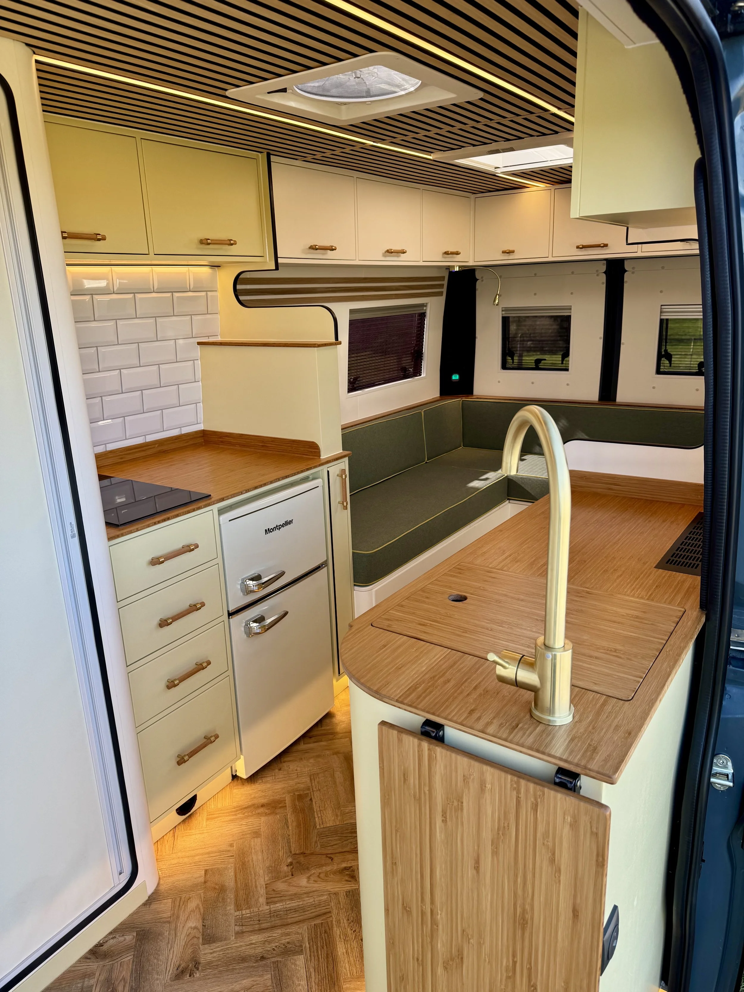 Interior of a small, modern camper van kitchen with cream cabinets, wooden countertops, and a green seating area with windows.