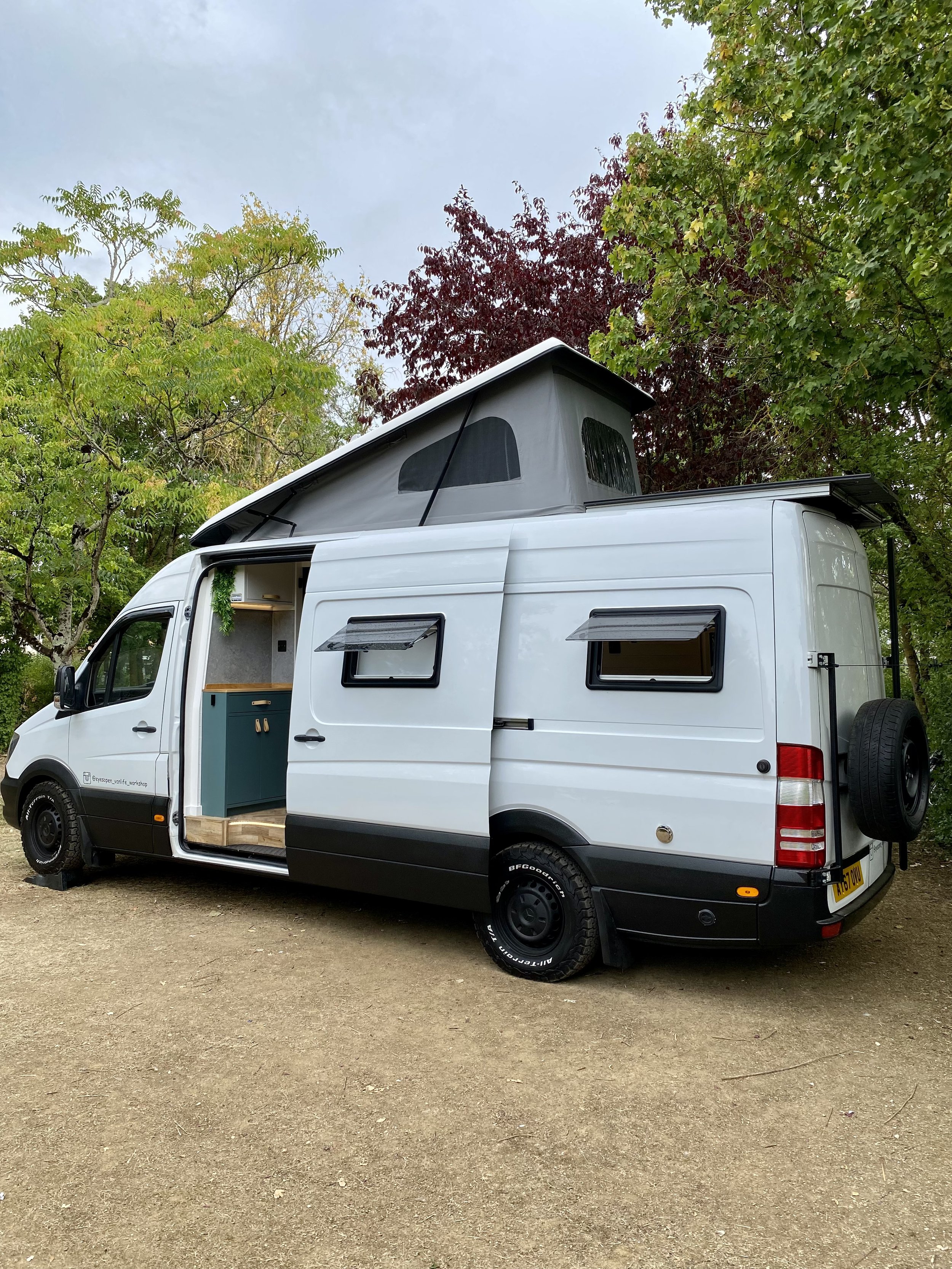 White camper van with a raised pop-up roof parked on dirt ground, surrounded by green and red trees.