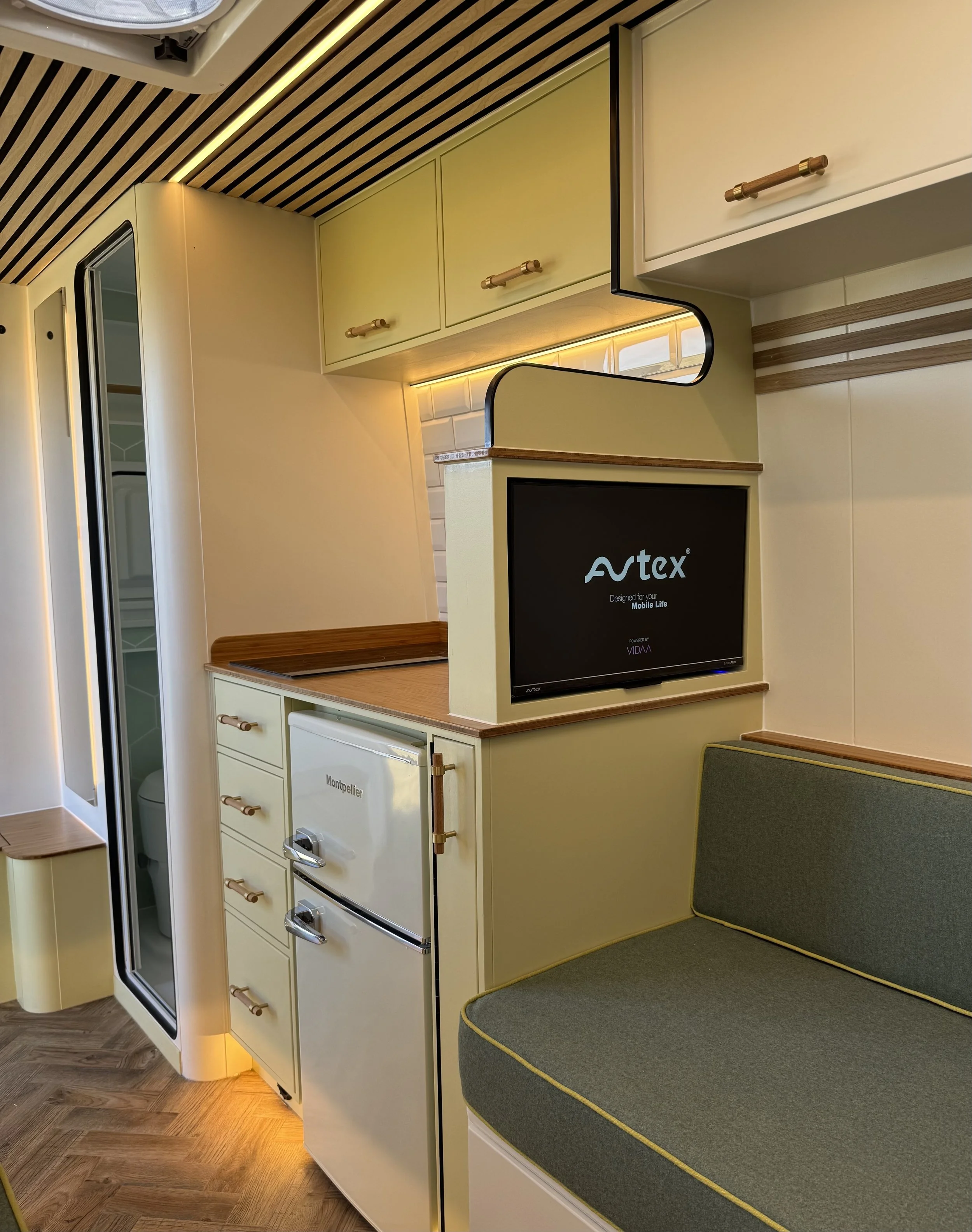 Interior of a modern space with shelves and a side table, featuring a black TV with the logo 'Avtex' on the screen, a green cushioned seating area, and a slatted wooden ceiling.