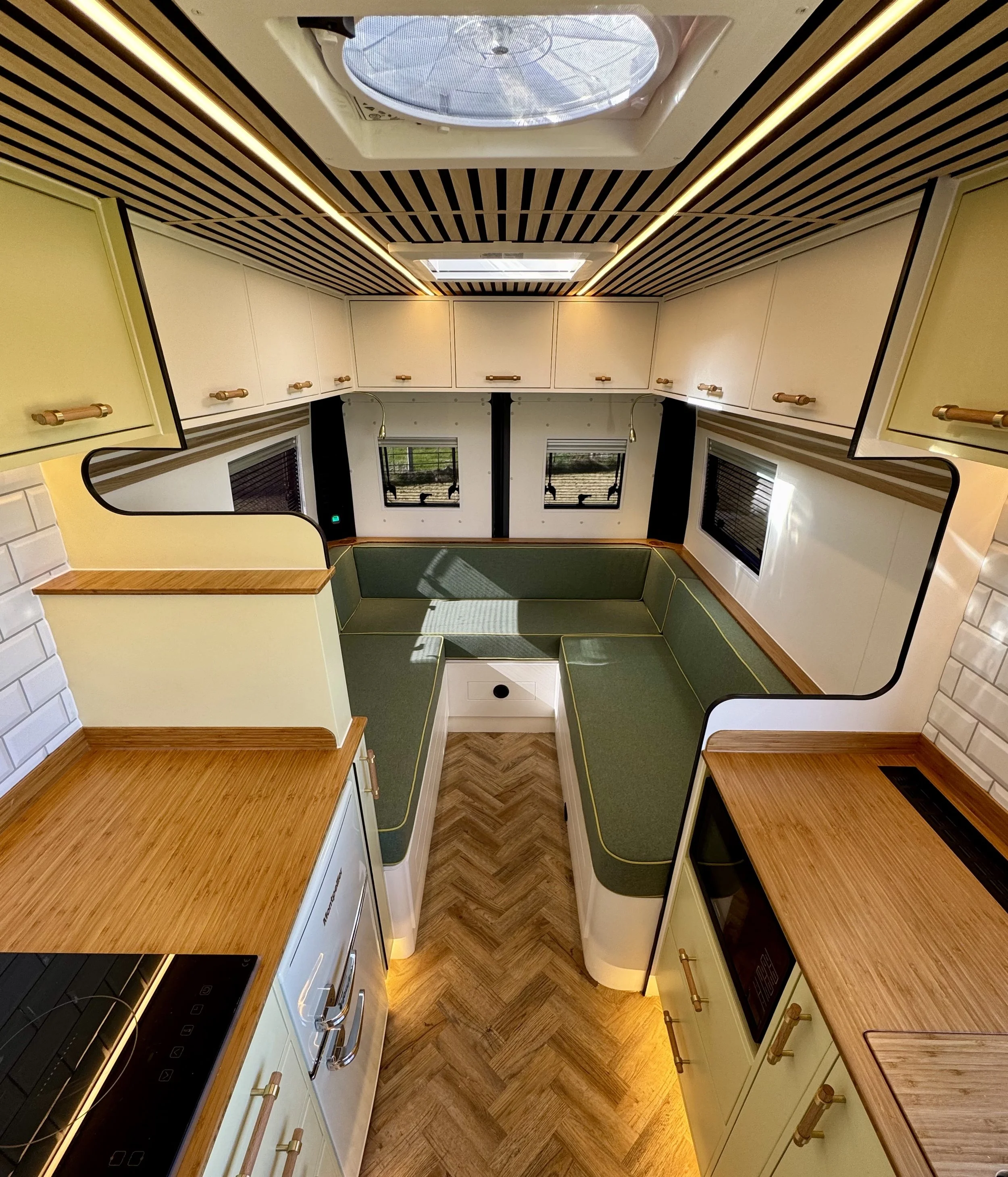 A cozy kitchen with wooden countertops, off-white cabinets with wooden handles, a stove, and three small windows above the countertops, with a ceiling featuring striped wood paneling and skylights.