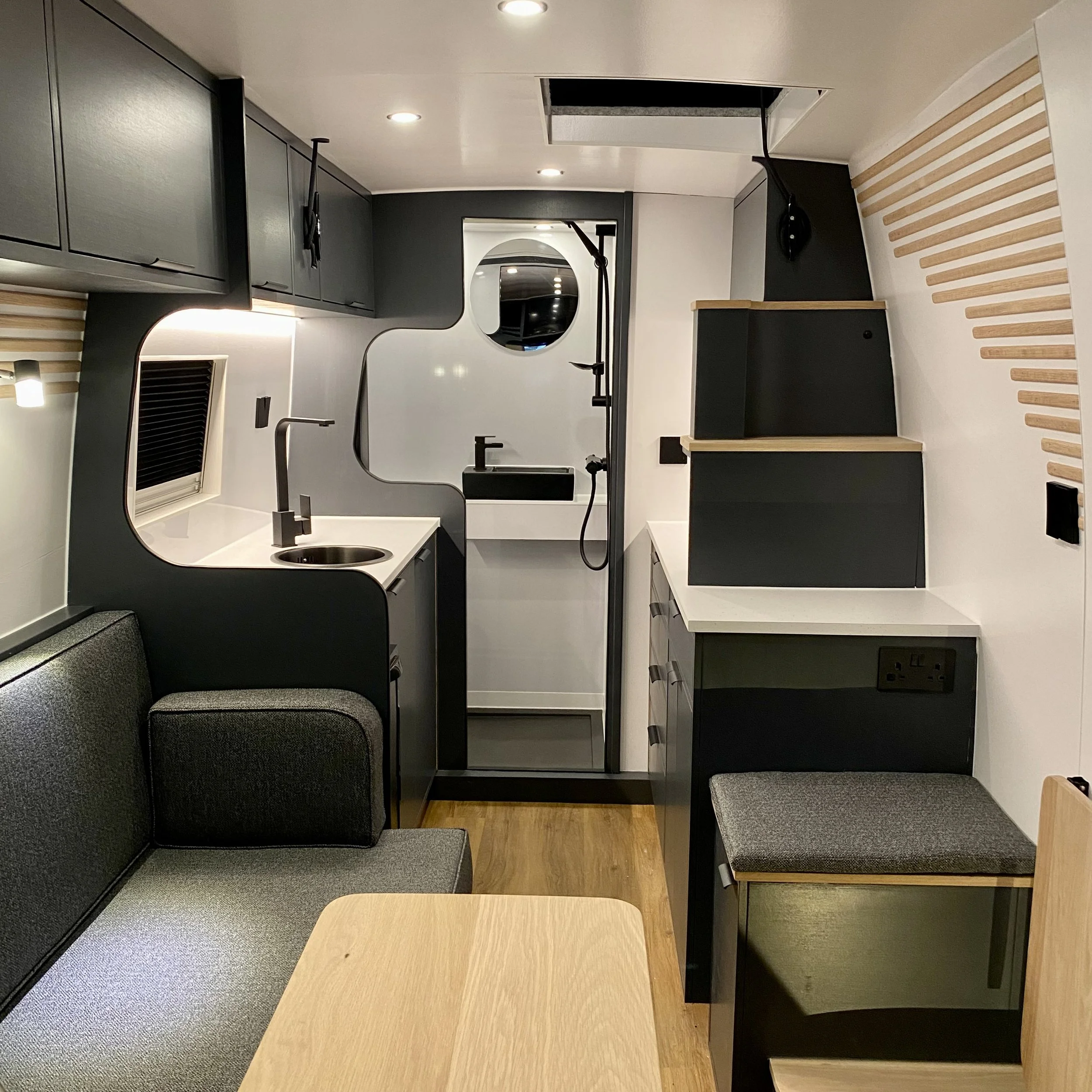 Modern interior space with seating area, black cabinets, and a mirror with an oval shape. Light-colored wood tables, black accents, and a mix of carpeted and wooden flooring. Ceiling has recessed lighting and an open ceiling design with a black pipe.