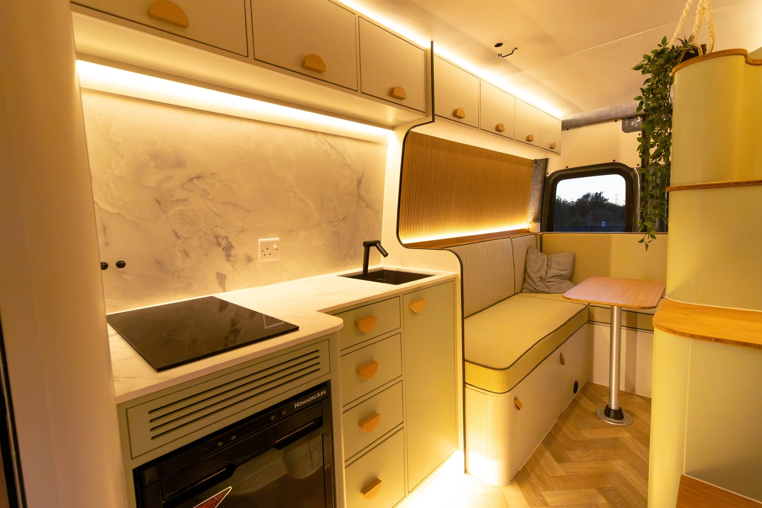 Small kitchenette with cabinets and a countertop. A black induction cooktop is visible on the counter, and there's a small sink nearby. The cabinetry has beige doors with tan handles. A cozy seating area with a yellow cushioned bench and a small wooden table is adjacent to the kitchenette, with a window and some green plants on the right side. The walls feature a marble-like texture and ambient lighting provides a warm glow.
