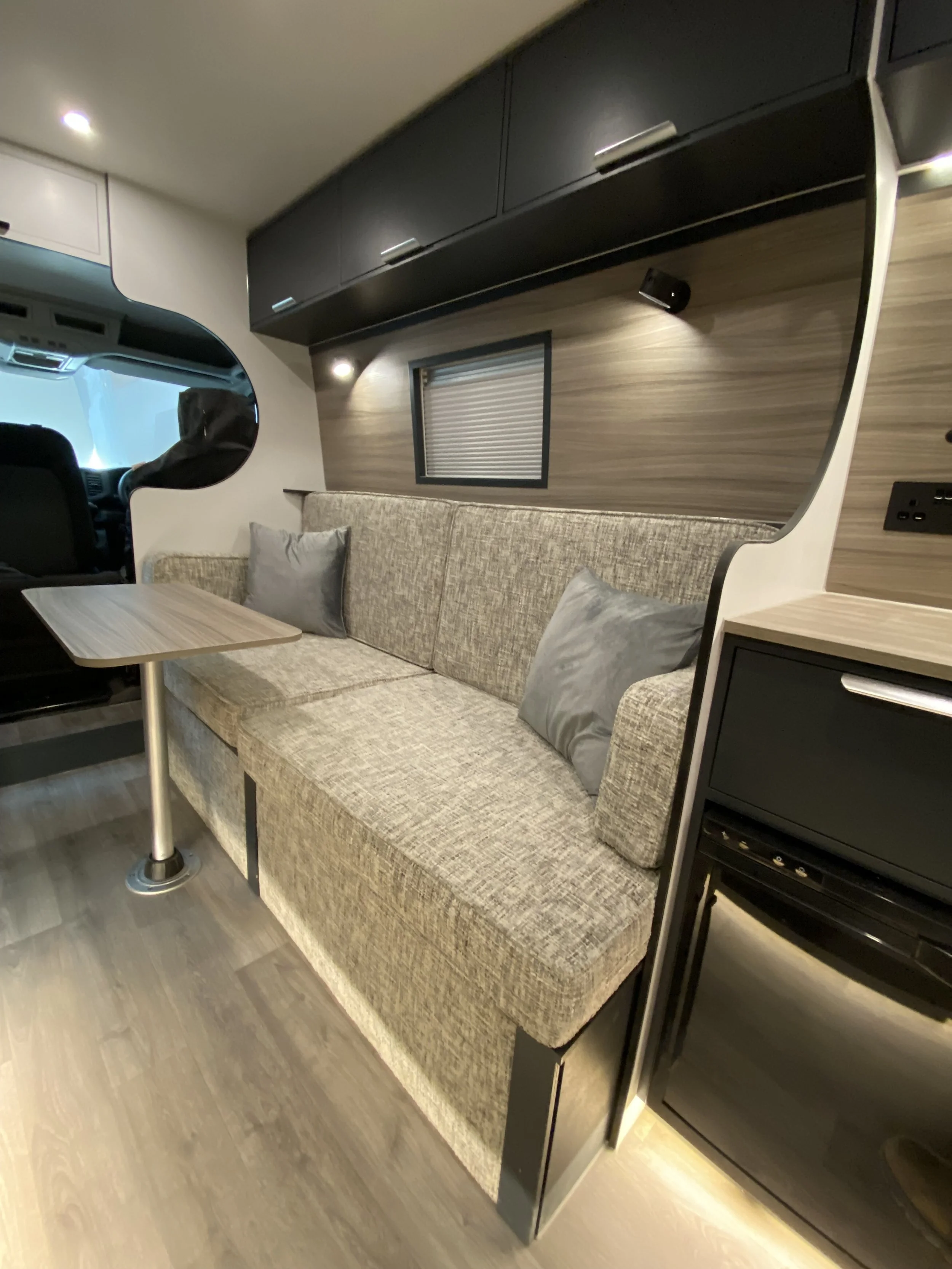 Interior of a modern camper or RV with a beige sofa, pillows, a small table, overhead black cabinets, and a window with closed blinds.