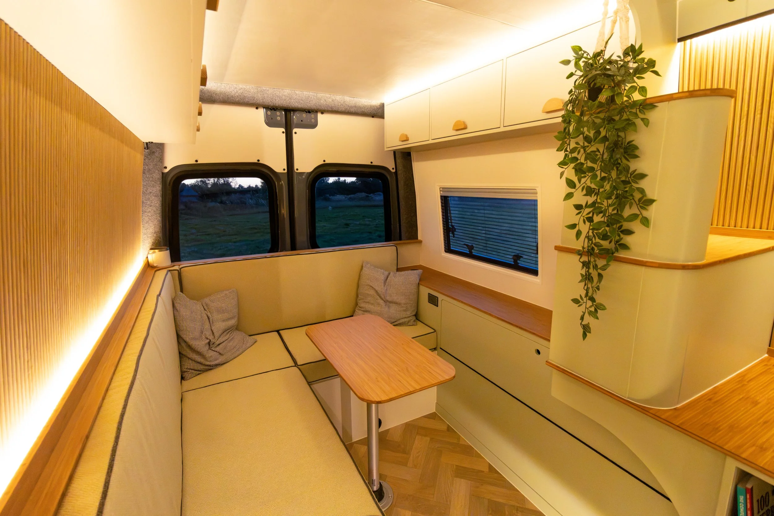 Interior of a cozy camper van with a corner seating area, small wooden table, large windows with blinds, wooden cabinets, and a climbing plant on the wall, illuminated with warm lighting.