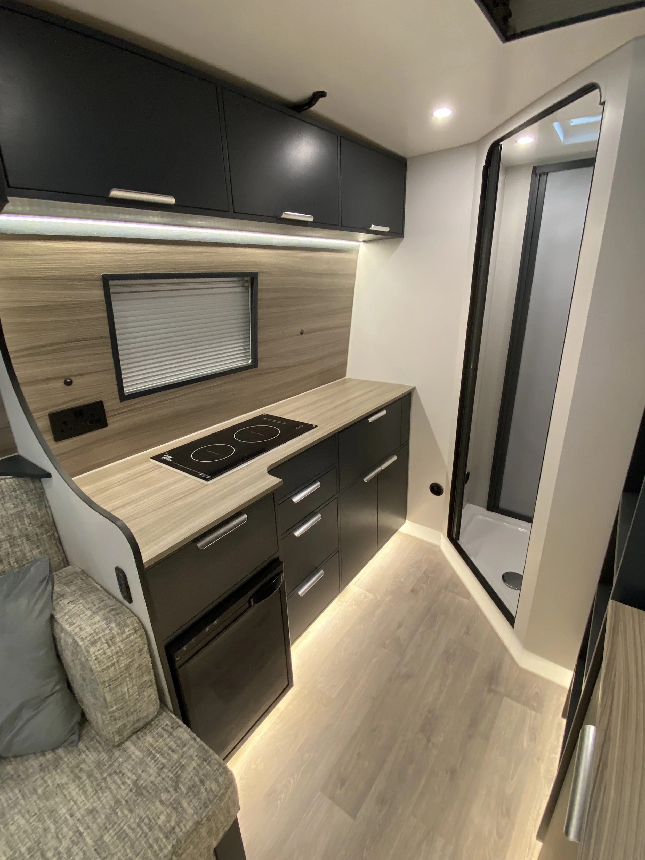 Interior of a compact kitchen in a mobile home or RV with dark cabinets, a woodgrain backsplash, a small cooktop, and a shower stall in a small bathroom area.