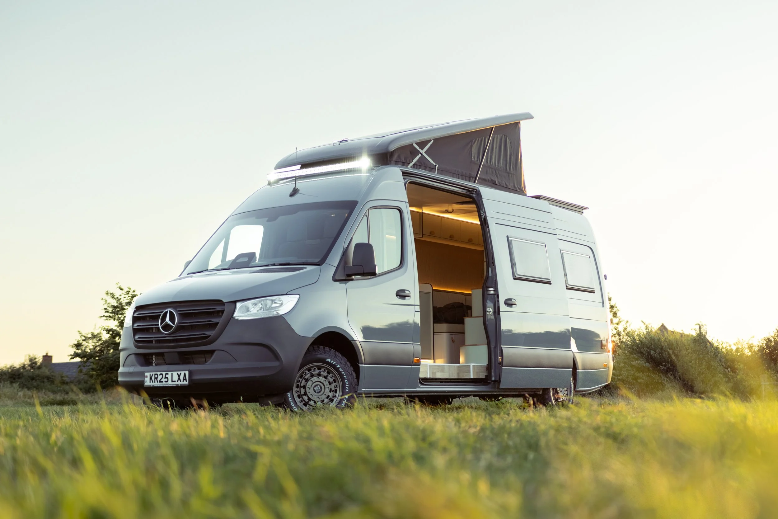 A white camper van with an open door, parked in a grassy field during sunset, with a pop-up roof raised.