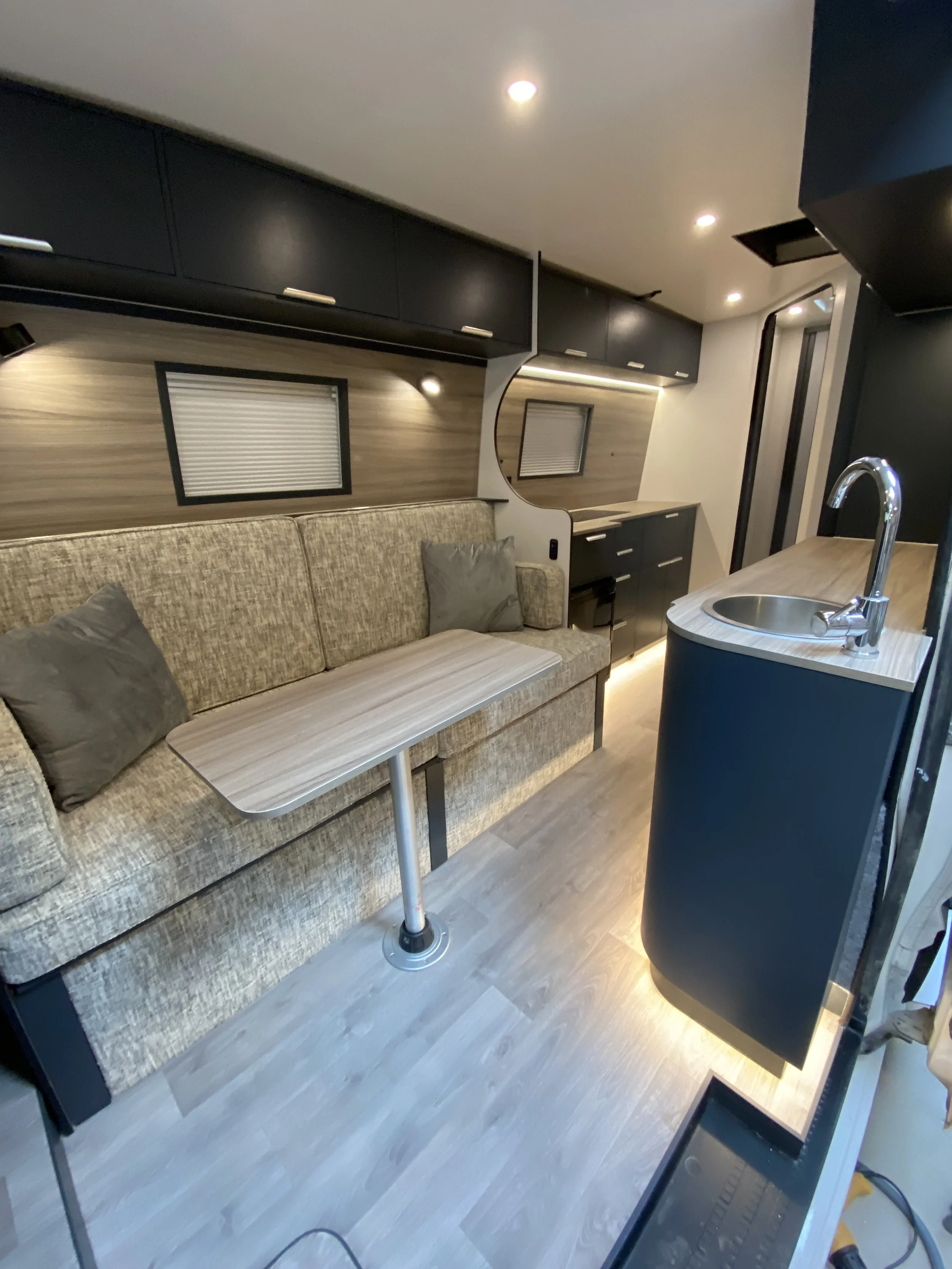 Interior of a modern recreational vehicle or tiny home with a beige couch, small black table, kitchen sink, upper and lower black cabinets, wood paneling, and small windows, under ceiling lights.