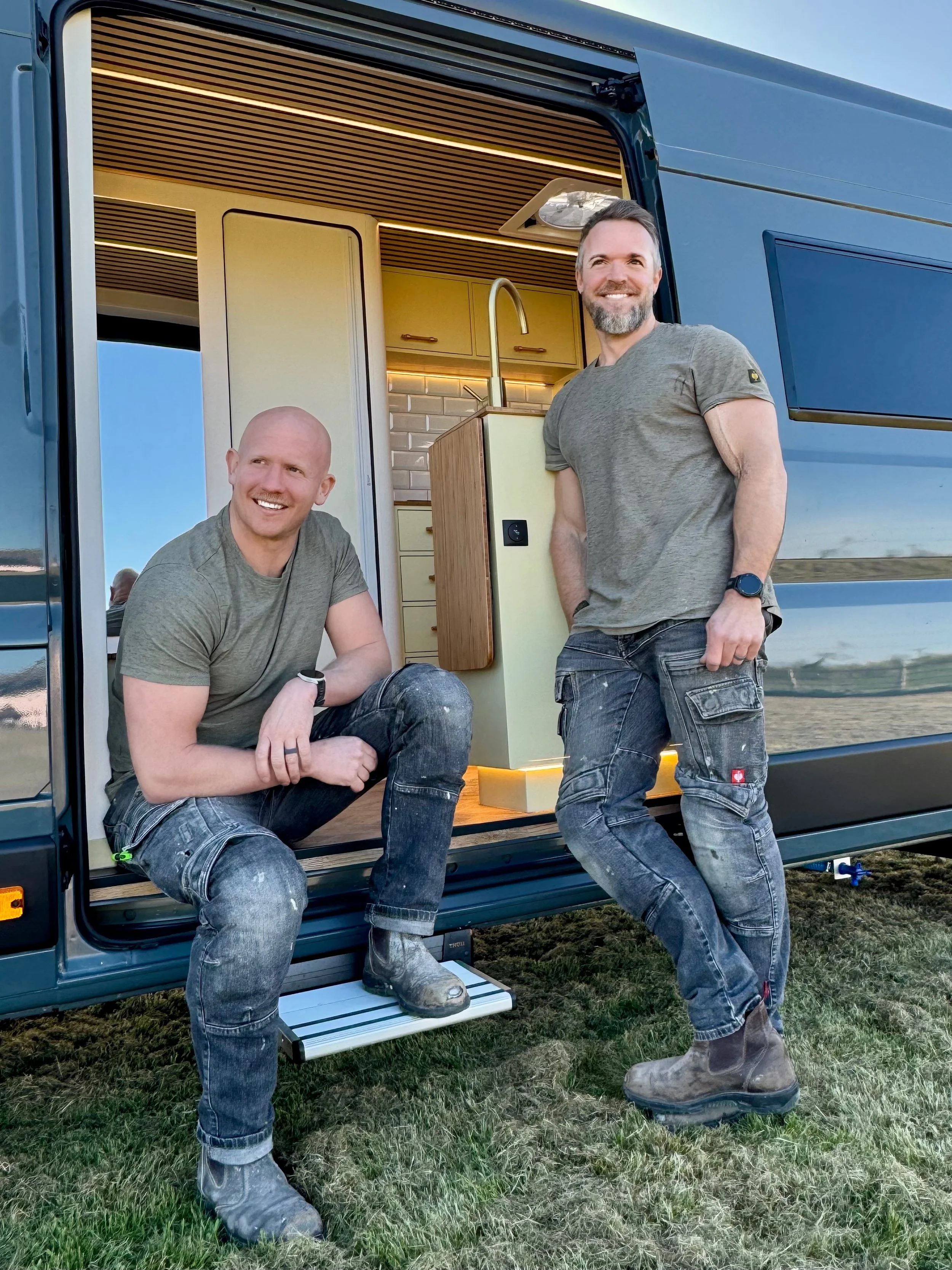Two men with gray hair and beards posing outside a mobile home, one sitting and one standing, both wearing gray t-shirts, jeans, and work boots, with a small kitchen visible inside the mobile home.
